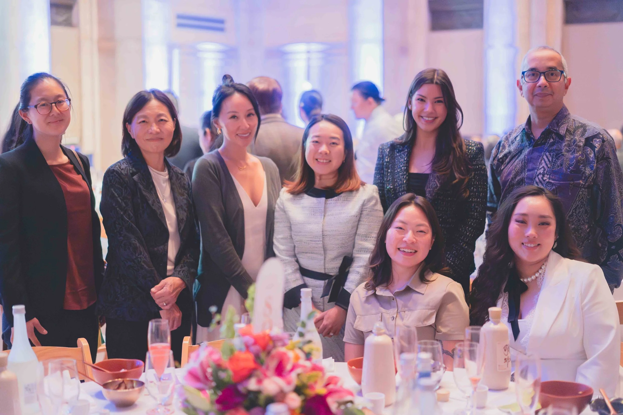 A group of nine women and one man posing at a formal event, with a decorated table featuring flowers and beverages in the foreground. They are smiling and dressed in business or semi-formal attire, with colorful lighting in the background.