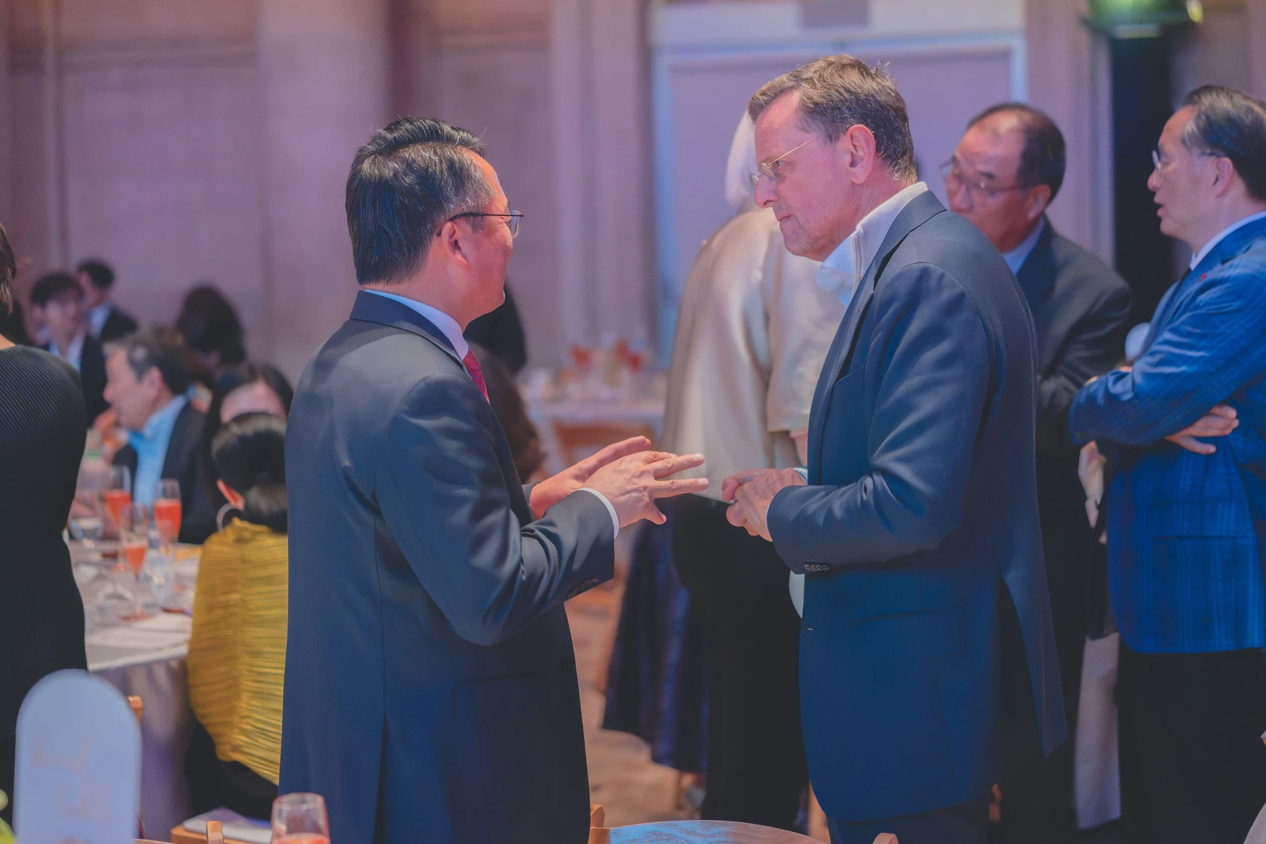 Two men in suits are engaged in a conversation at a formal event, with other attendees in the background. They appear to be in a restaurant or banquet hall.