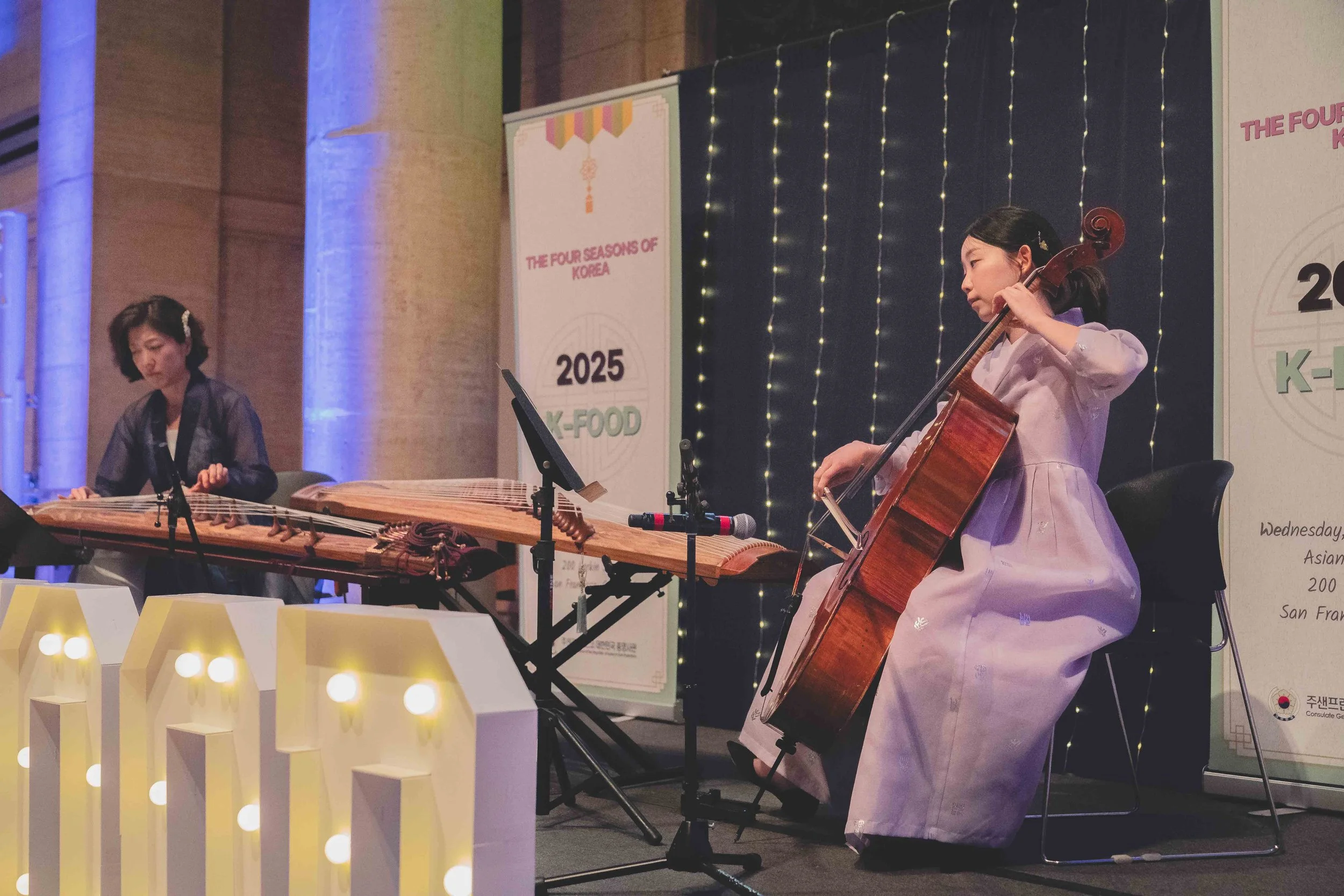 Two young girls performing traditional Korean music at an indoor event. One girl is playing a gayageum, a traditional Korean stringed instrument, while the other girl is playing a cello. Behind them are banners with event details and decorative strin