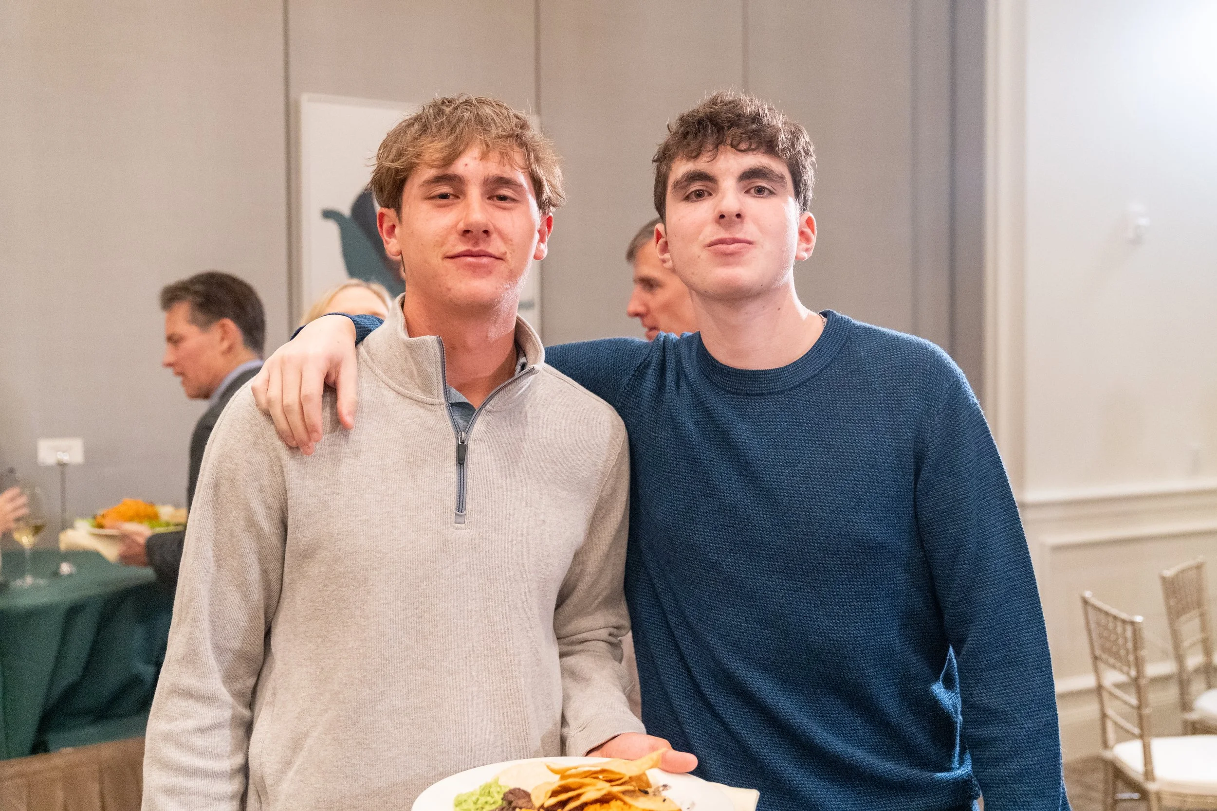 Two young men standing together, one with an arm around the other's shoulder, holding a plate of food with chips and guacamole at an indoor event. Several people and a table with food are in the background.