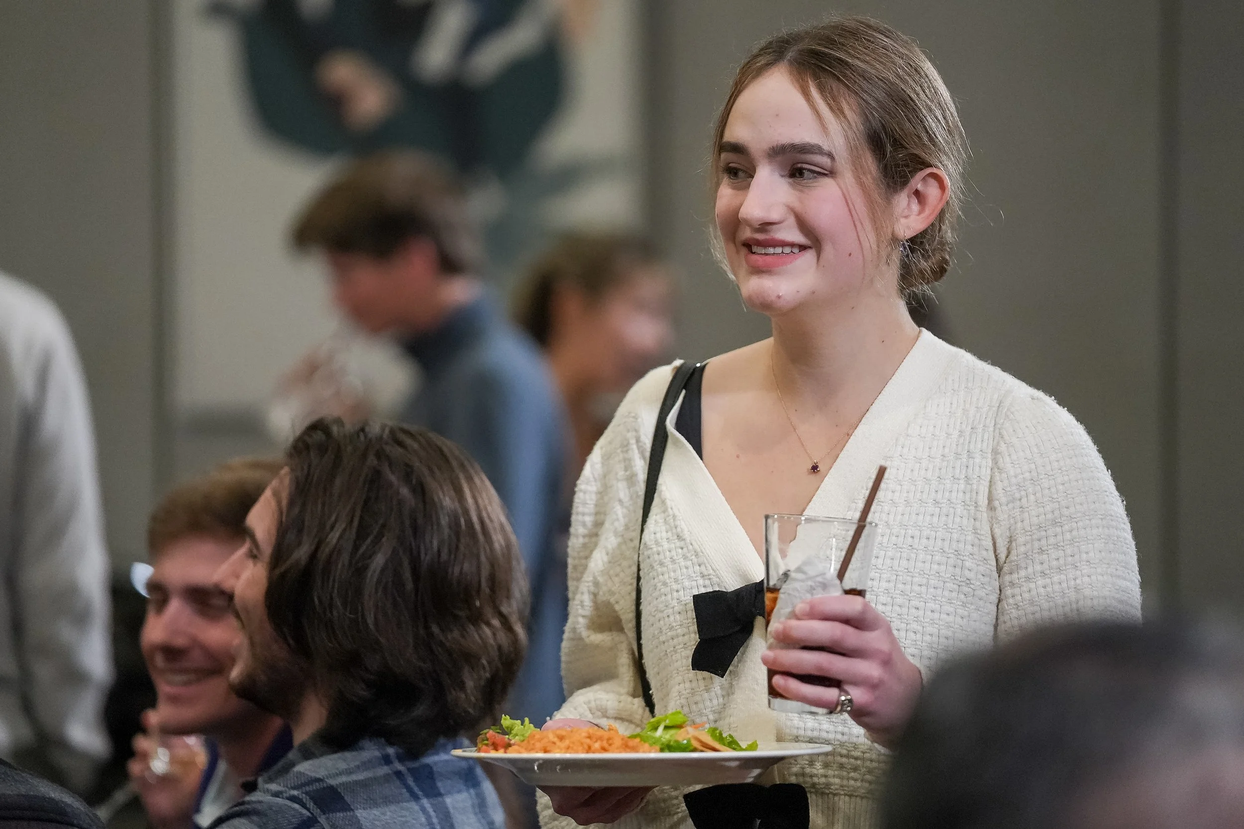 Woman holding a plate of food and a drink at a social event with people in the background.