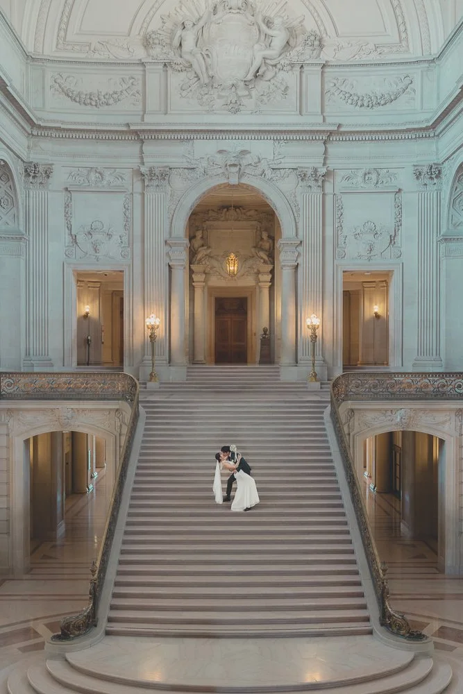 Two brides in wedding attire embracing on grand staircase inside ornate building.