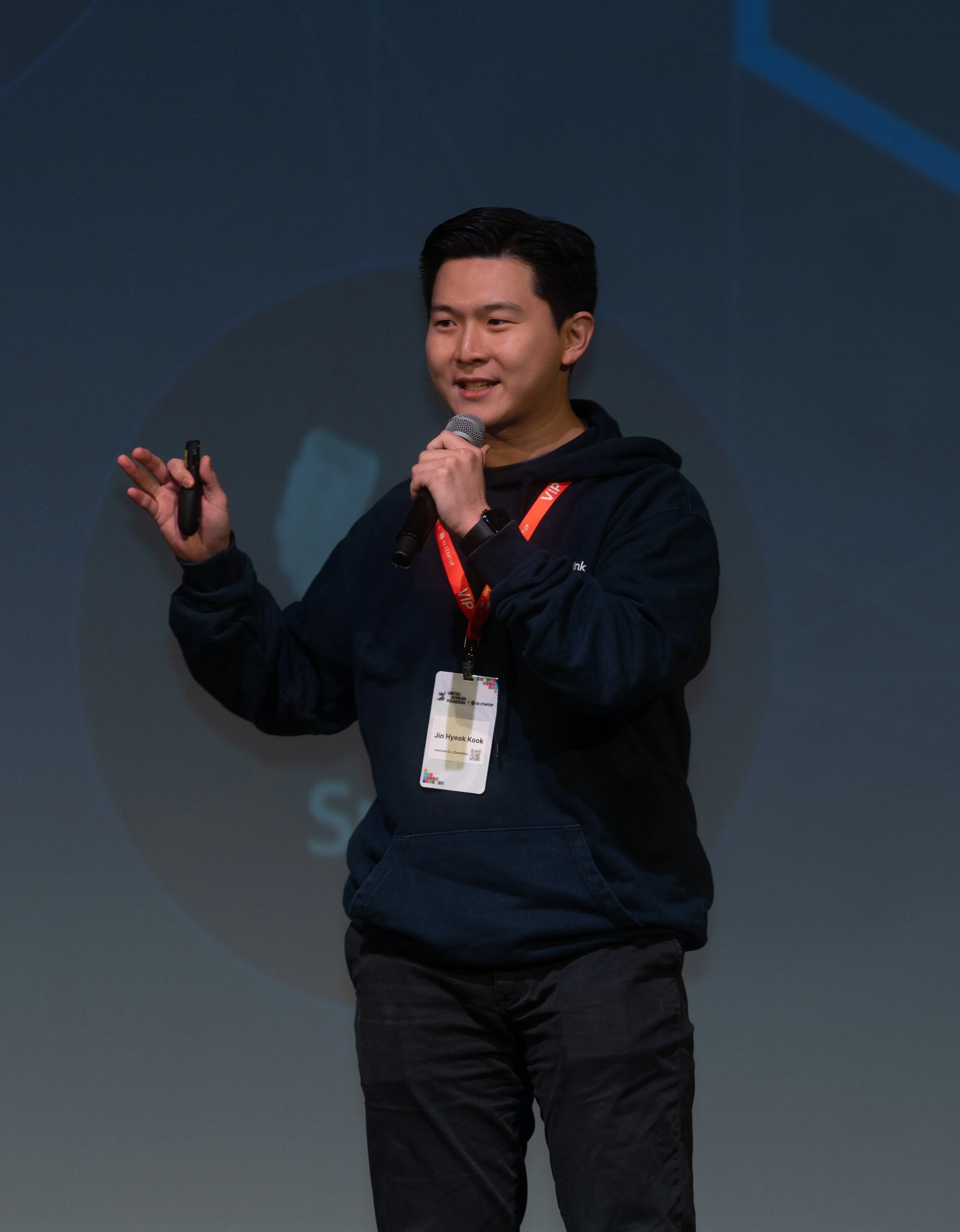 Person speaking on stage with a microphone, wearing a dark hoodie and a badge, and holding a remote control device.