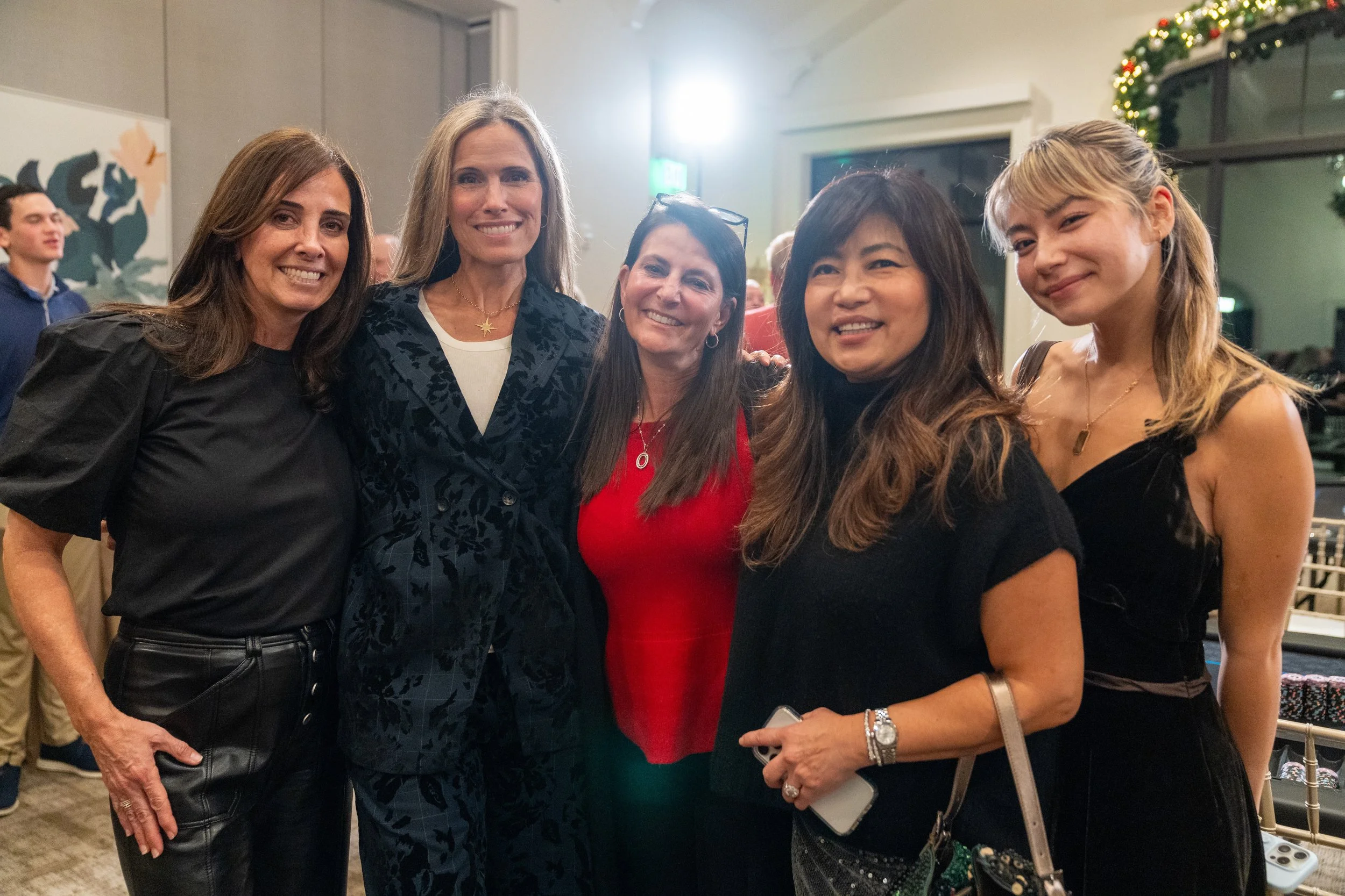 Group of five women smiling at a social gathering, one holding a smartphone, with holiday decorations in the background.