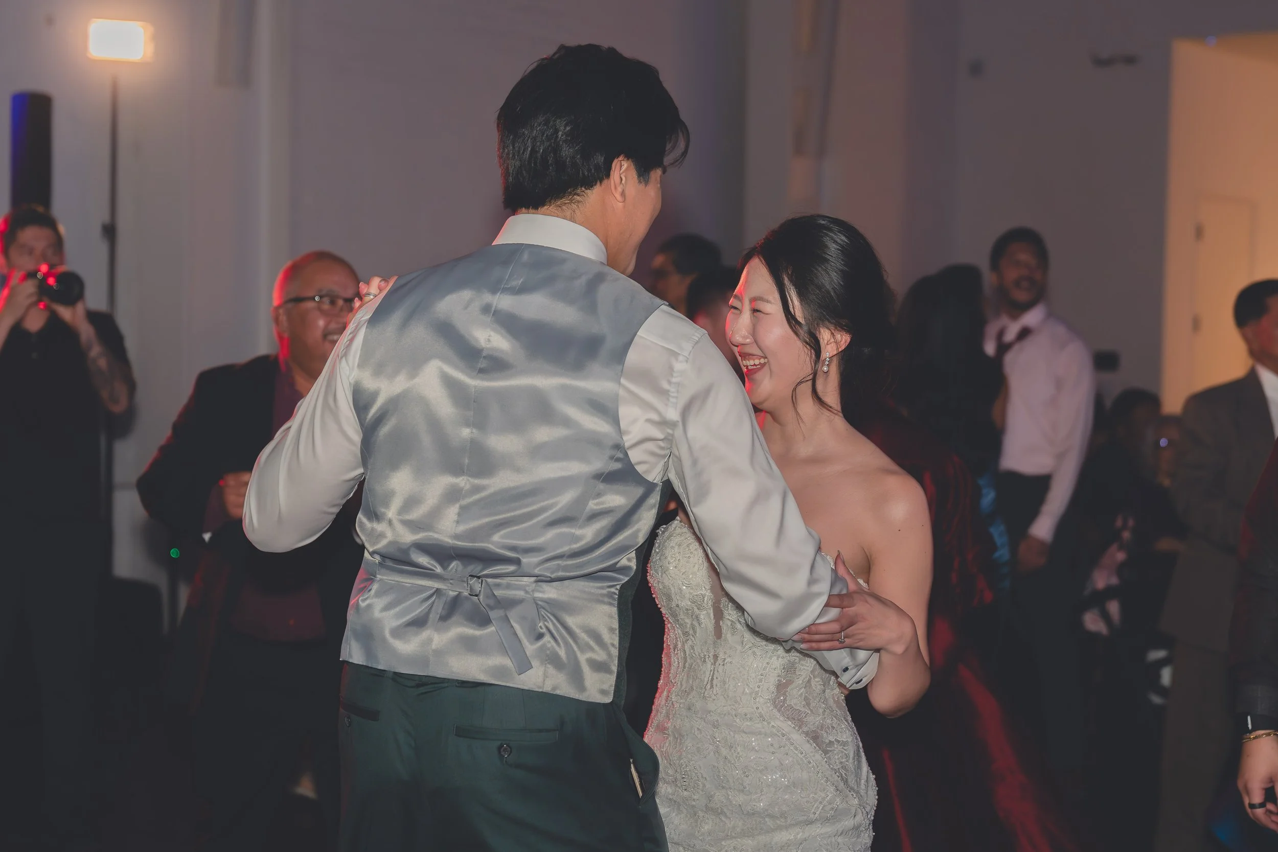 A couple dancing at a celebration, with the woman in a wedding dress and the man in a gray vest. People in the background are smiling, with one person taking a photo.