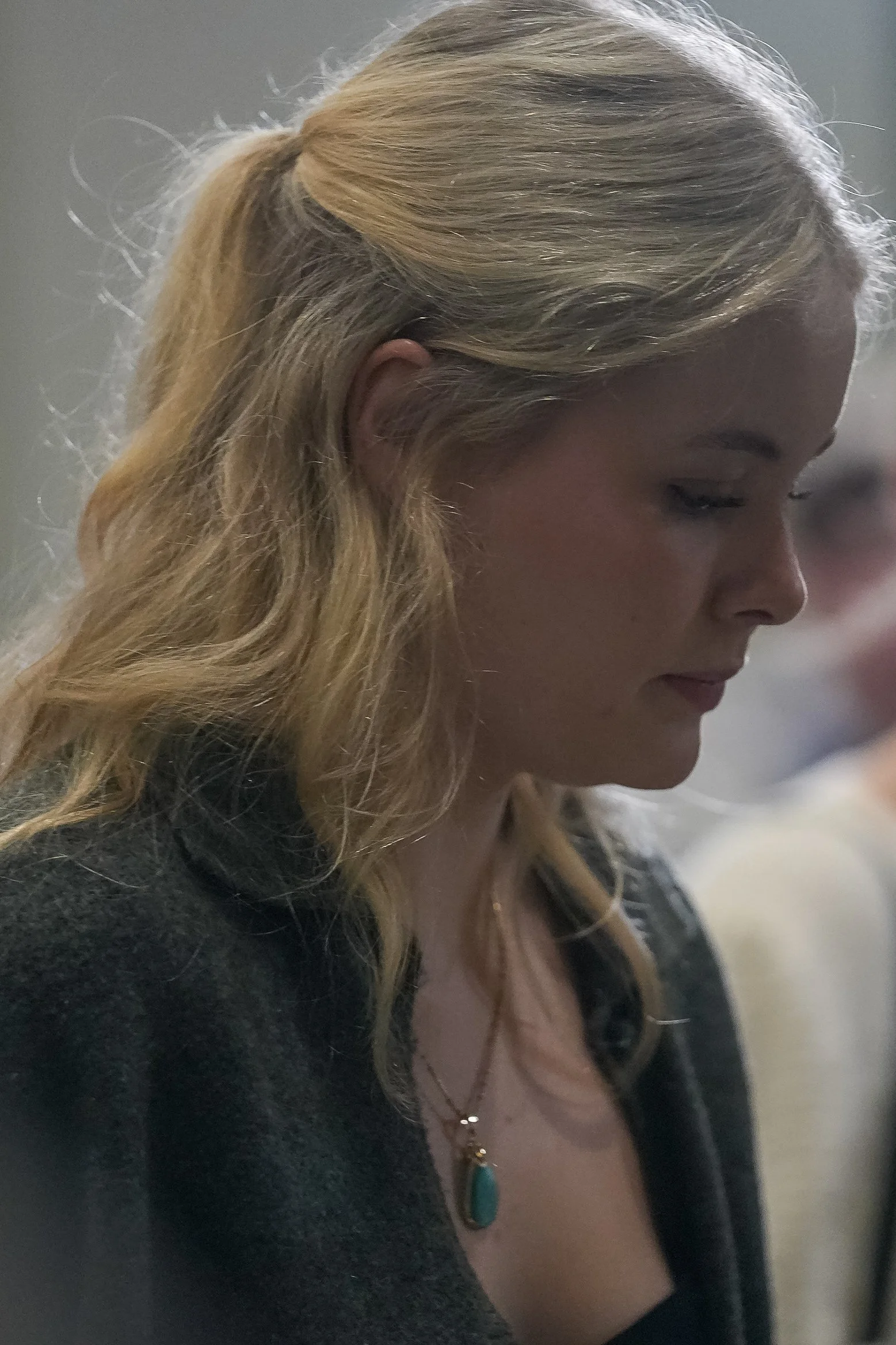 Side profile of a woman with blonde hair, wearing a dark top and necklace with a turquoise pendant.