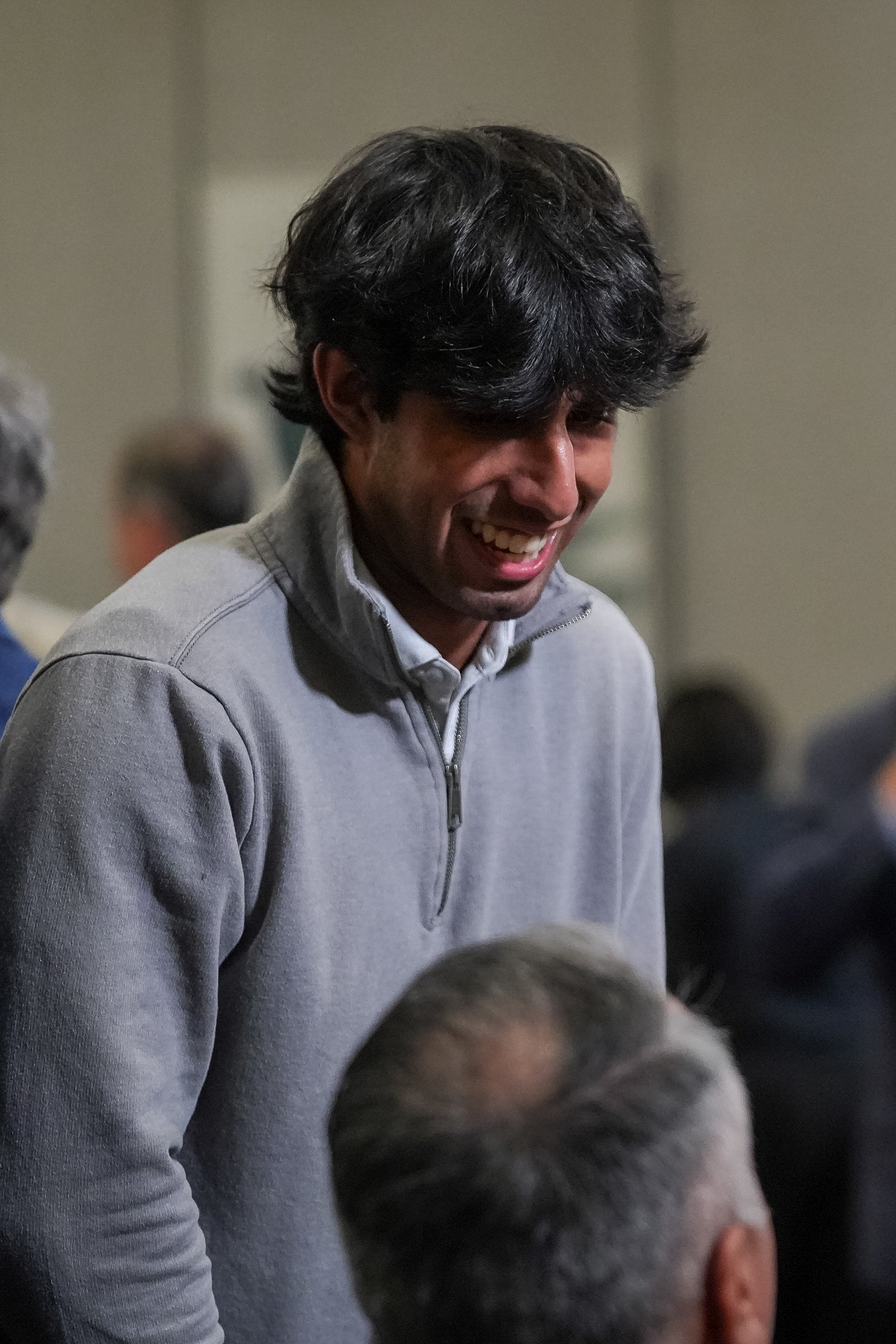 A person smiling, wearing a gray pullover, interacting with another individual seated.