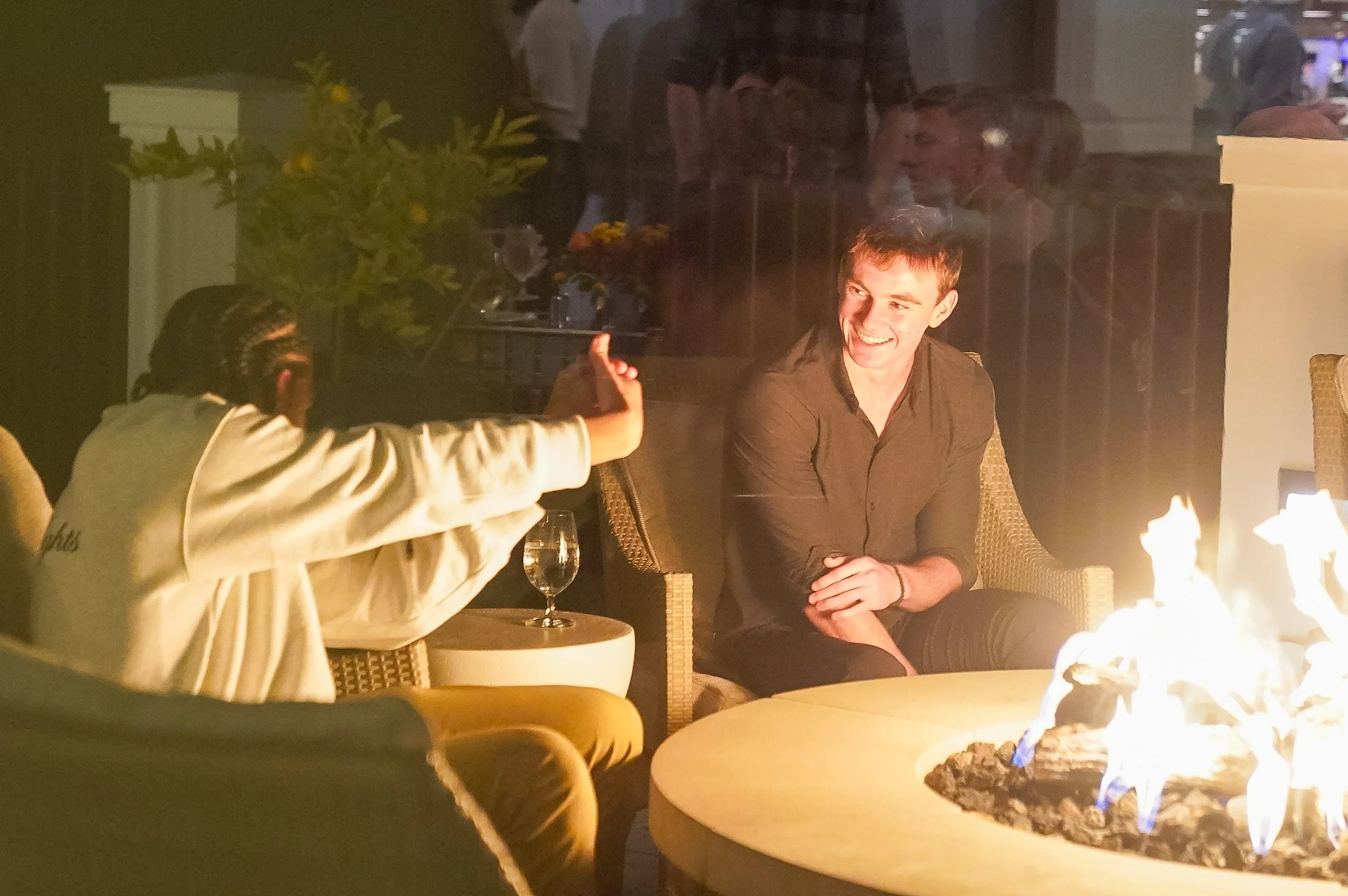 Two people sitting outdoors by a fire pit, one gesturing, the other smiling, with drinks on a nearby table.