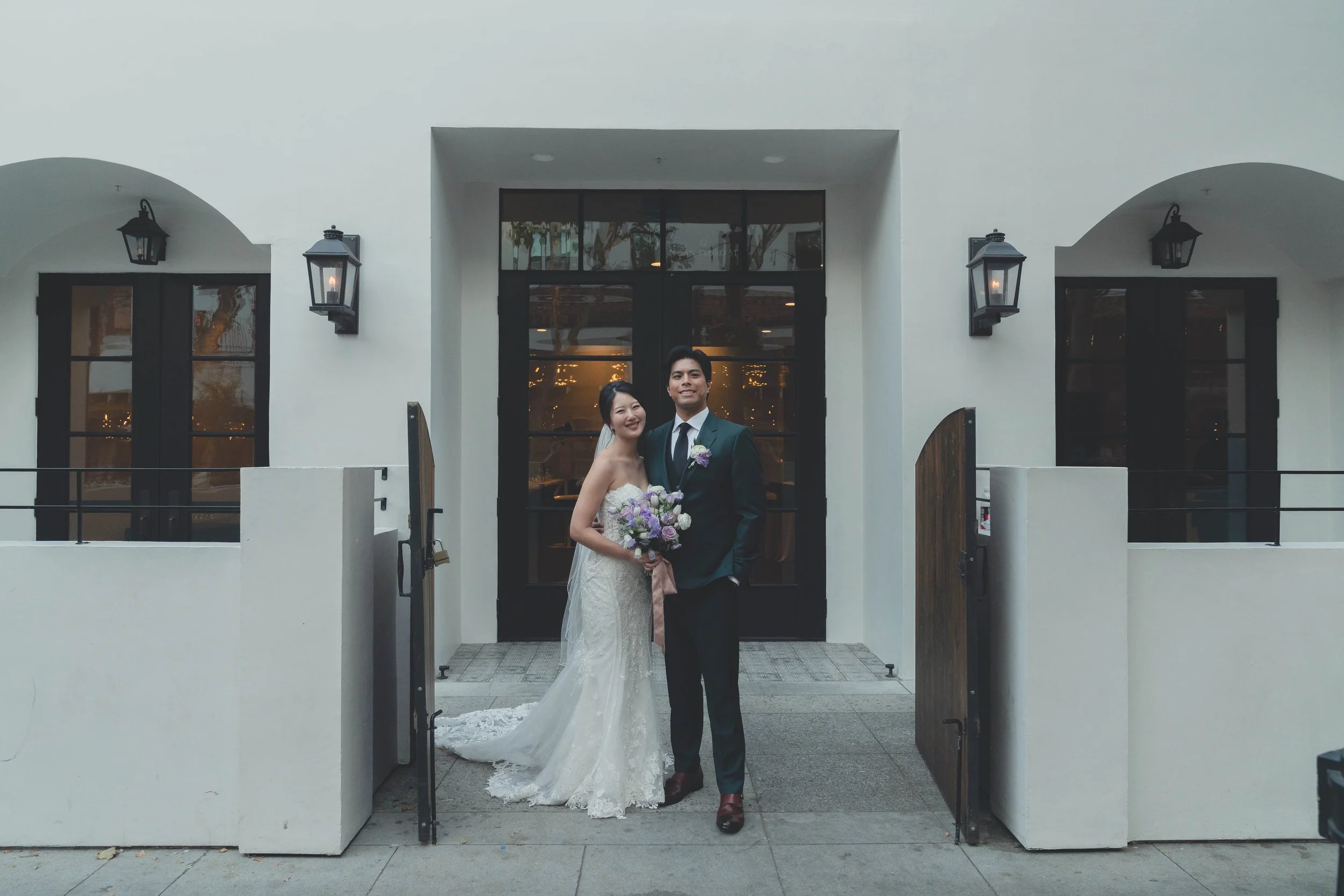 A newlywed couple standing outside in front of a building entrance, dressed in wedding attire. The bride is wearing a white gown and holding a bouquet of purple flowers, while the groom is in a black suit with a purple boutonniere. The background features white walls, black doors, and wall-mounted lanterns.