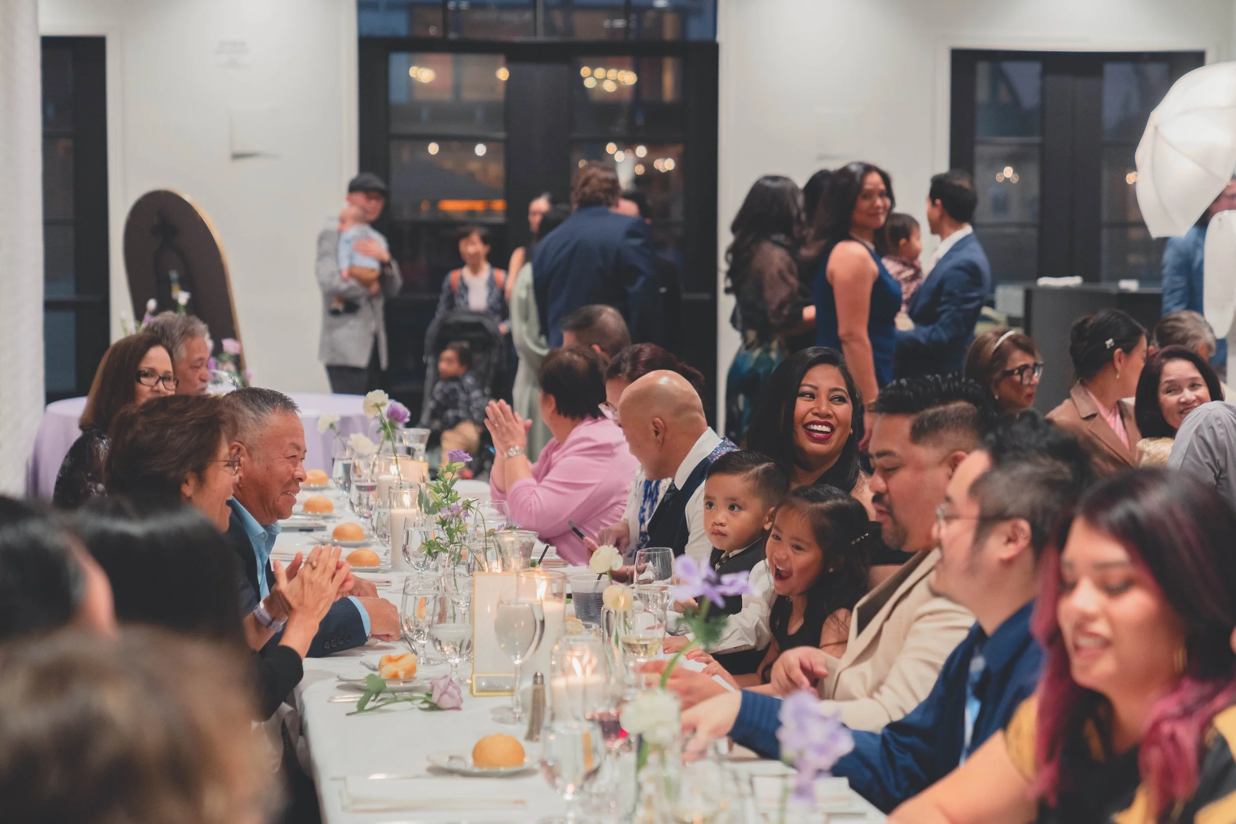 A lively group of people seated at a long table during an indoor gathering, likely a celebration. The table is elegantly set with flowers, candles, and glassware. People are engaged in conversation, smiling, and some are clapping.