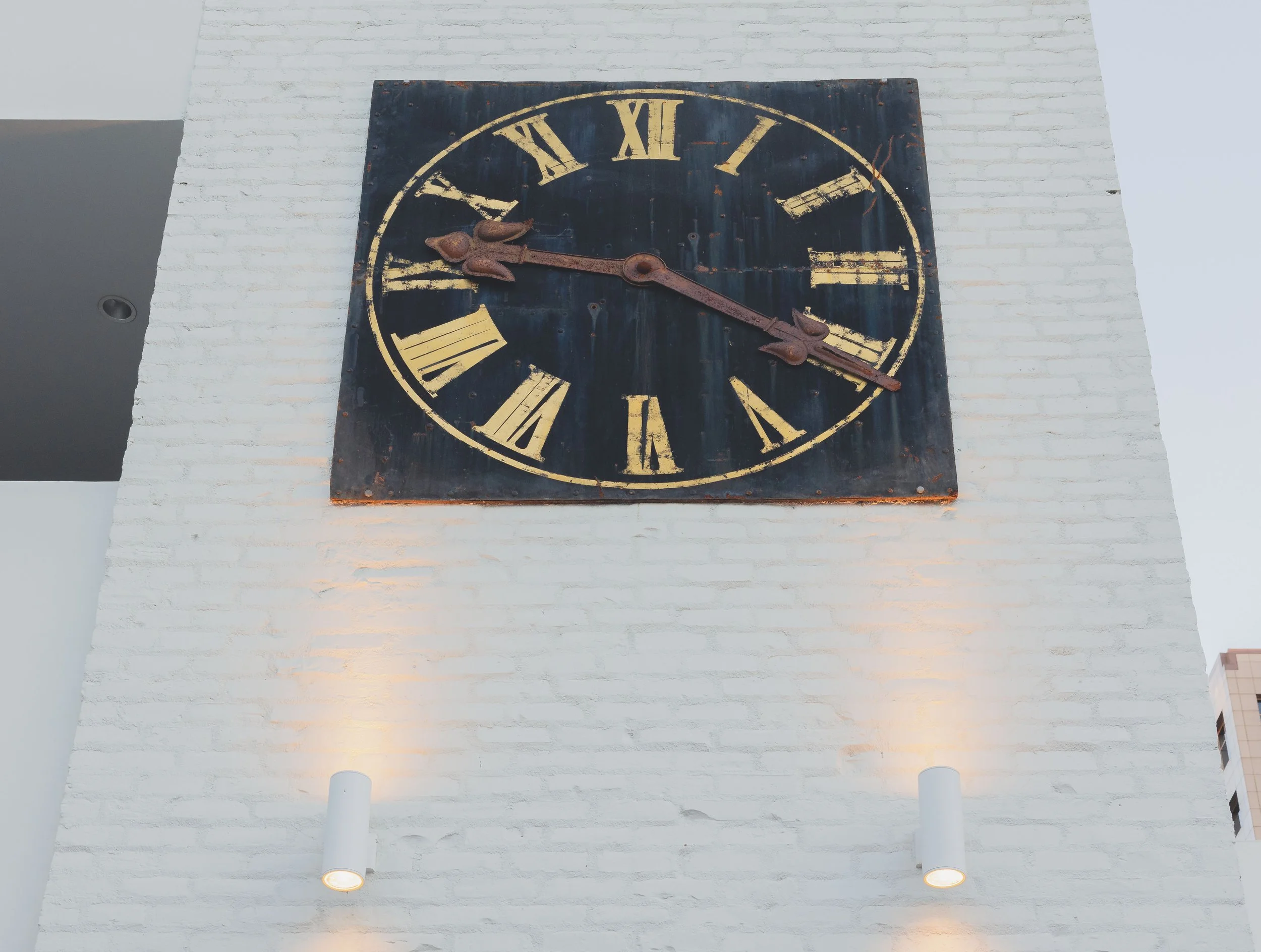 A vintage clock with Roman numerals on a white brick wall, accompanied by two cylindrical wall lights below it.