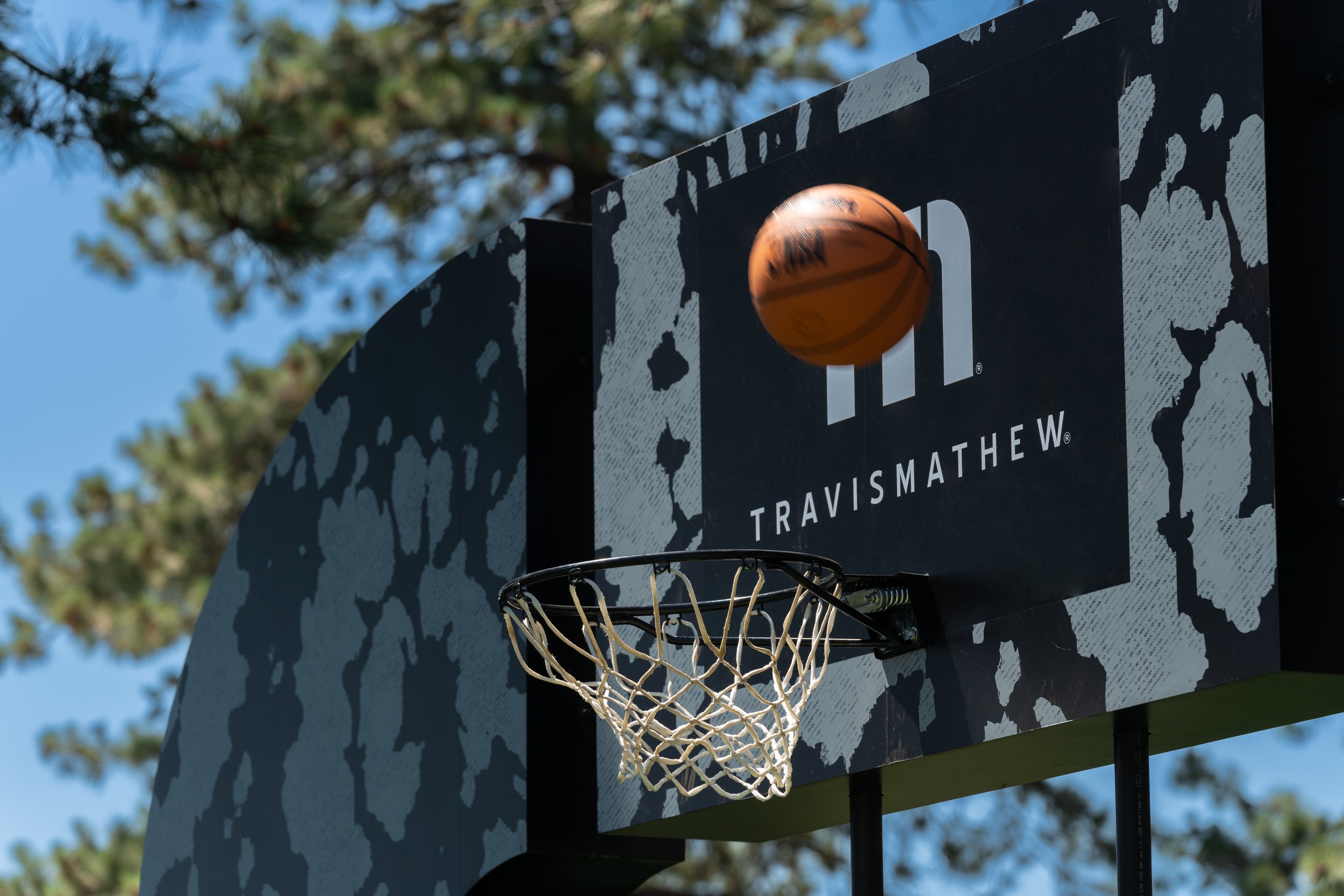 Basketball hoop with net, backboard featuring TravisMathew logo, and a basketball in mid-air against a tree-lined outdoor background.