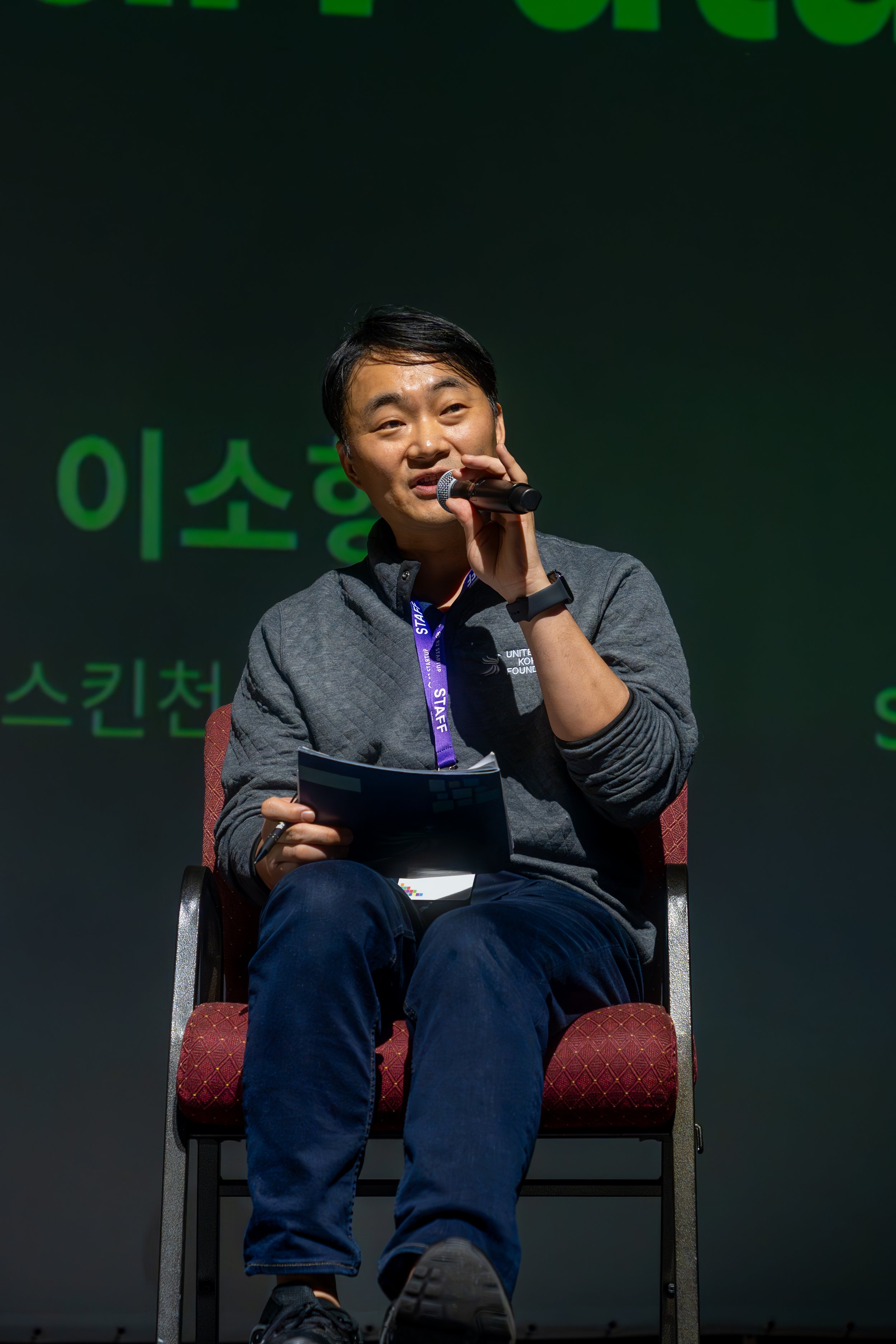 Person speaking into a microphone on stage, wearing a staff lanyard, seated in a chair, with green text in the background.