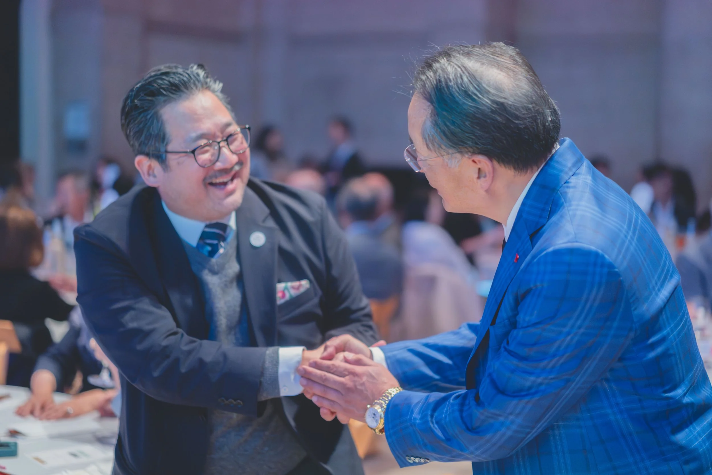 Two men shaking hands and smiling in a crowded, formal indoor setting, possibly at a conference or banquet.
