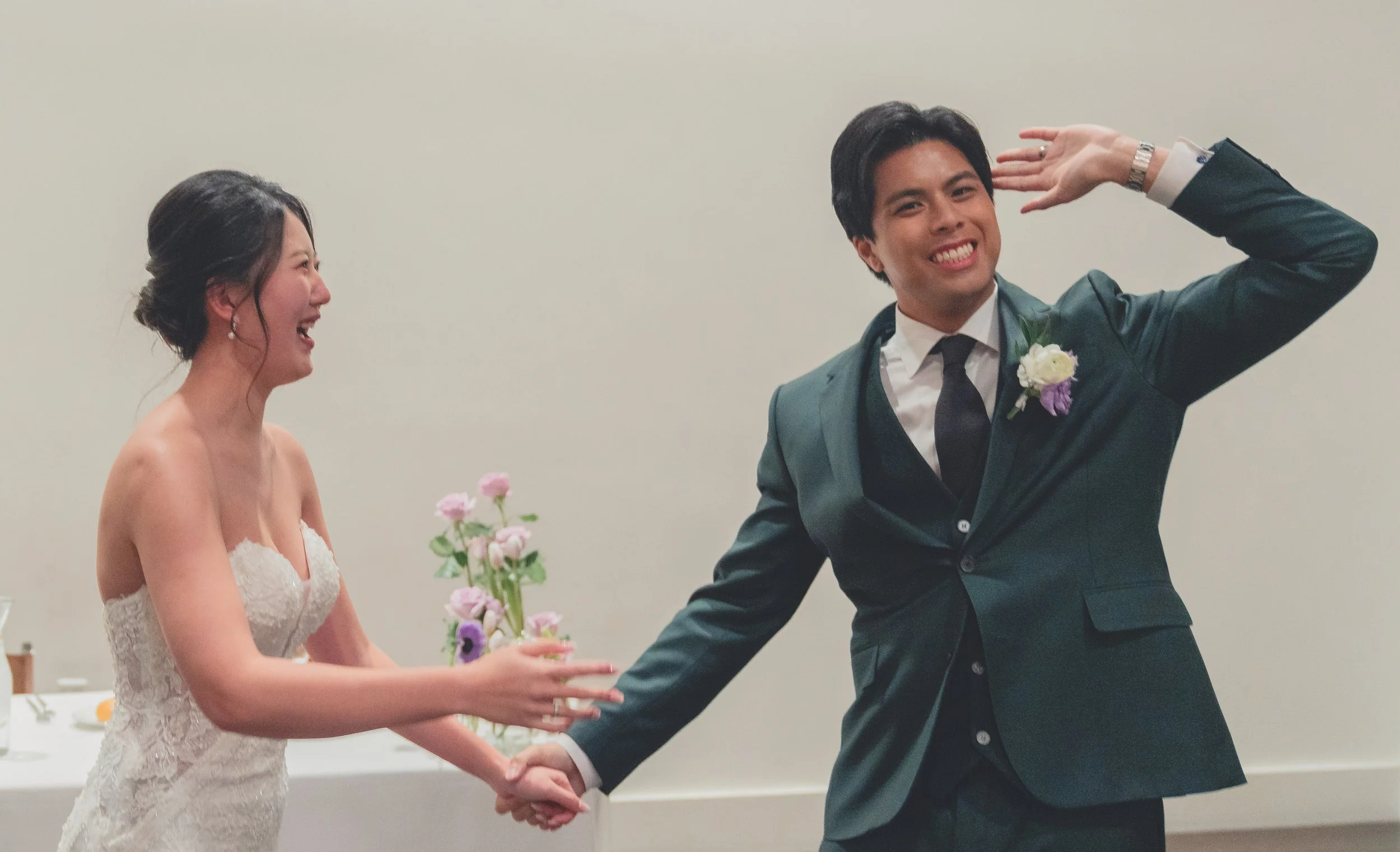 A bride and groom laughing and holding hands at a wedding reception, with the groom playfully posing. The bride is wearing a white lace dress and the groom is in a dark suit with a boutonniere. A table with flowers is in the background.