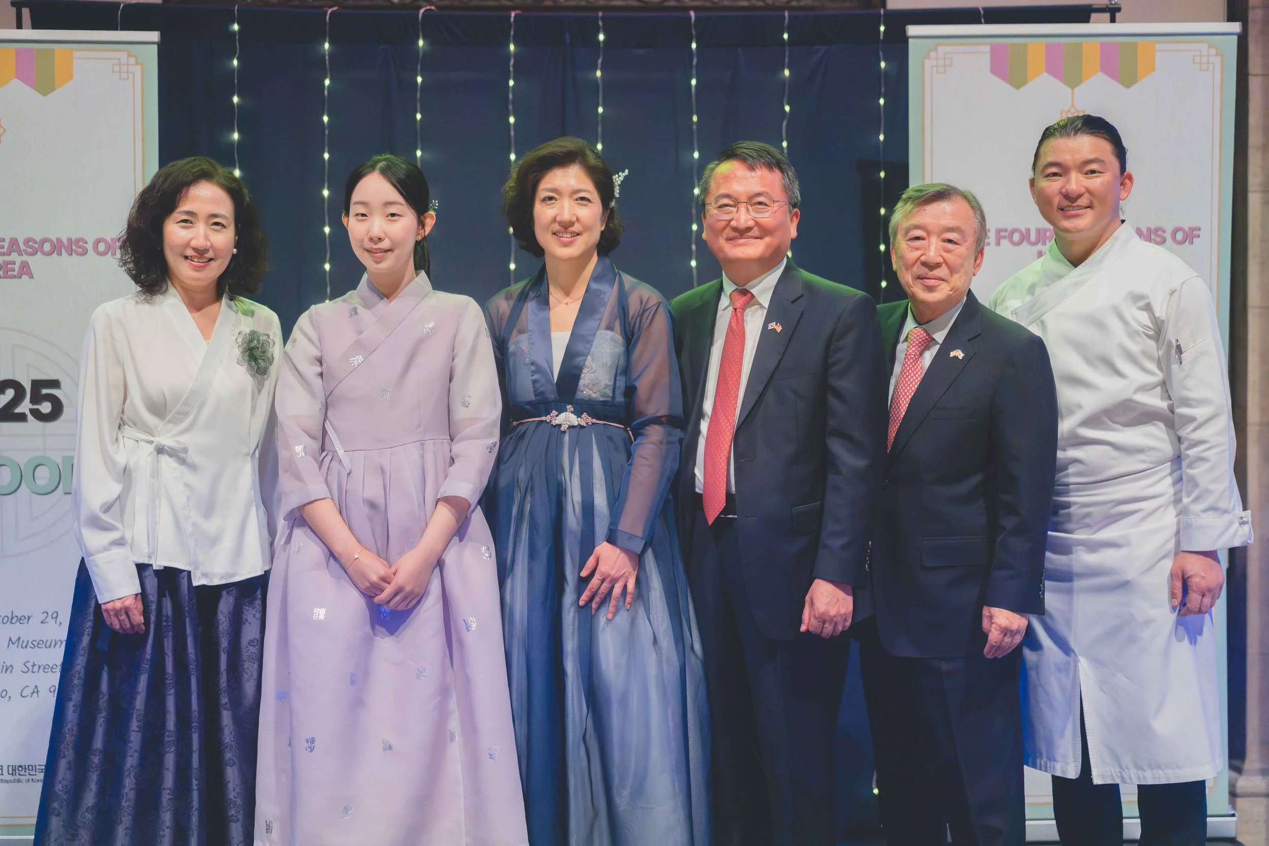 Six people in formal attire, three women in traditional Korean clothing and three men in suits, posing together at an event with a decorated backdrop.