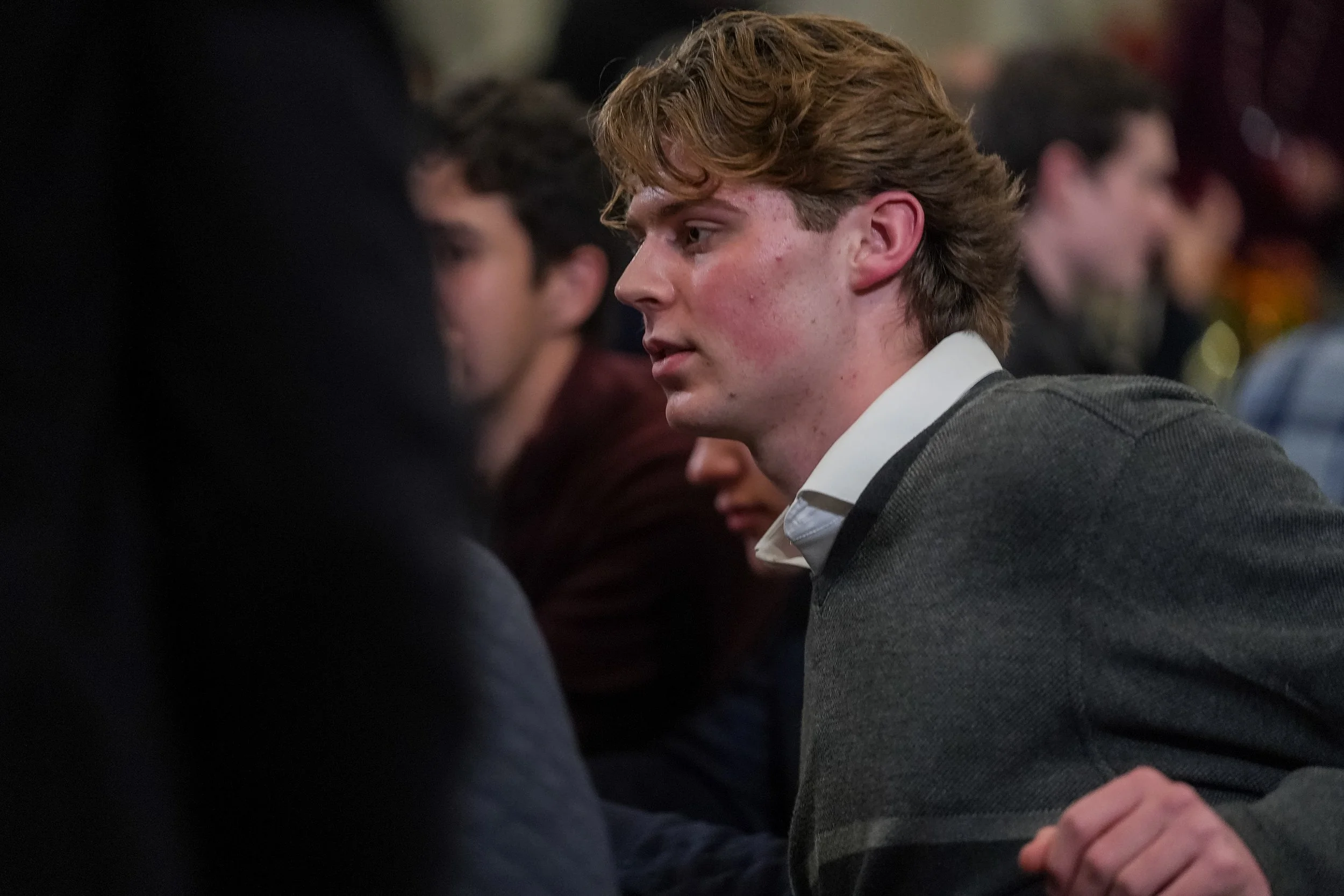 A young man with light brown hair in a gray sweater, sitting indoors among a group of people.