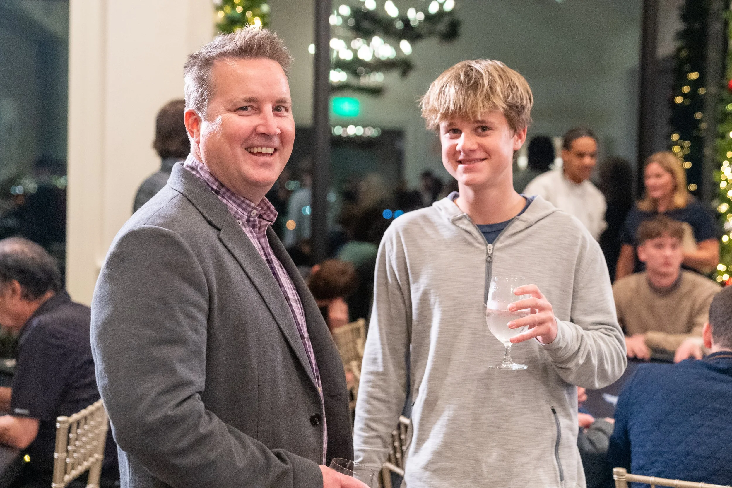 Two people smiling at a social gathering, one holding a drink, with holiday decorations in the background.