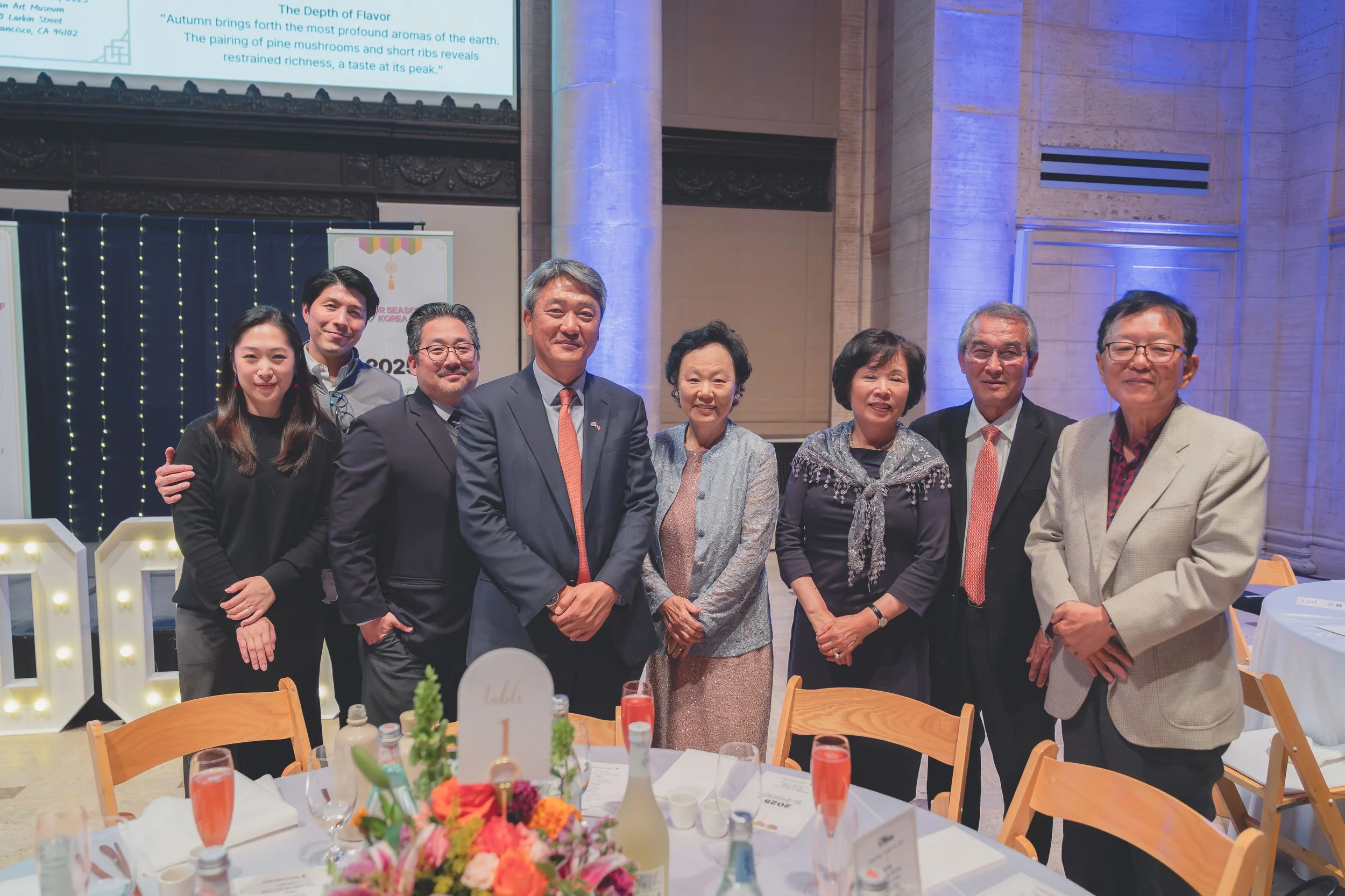 A group of nine people, both men and women, are standing together at a formal event inside a decorated venue with table settings and floral centerpieces, smiling at the camera.