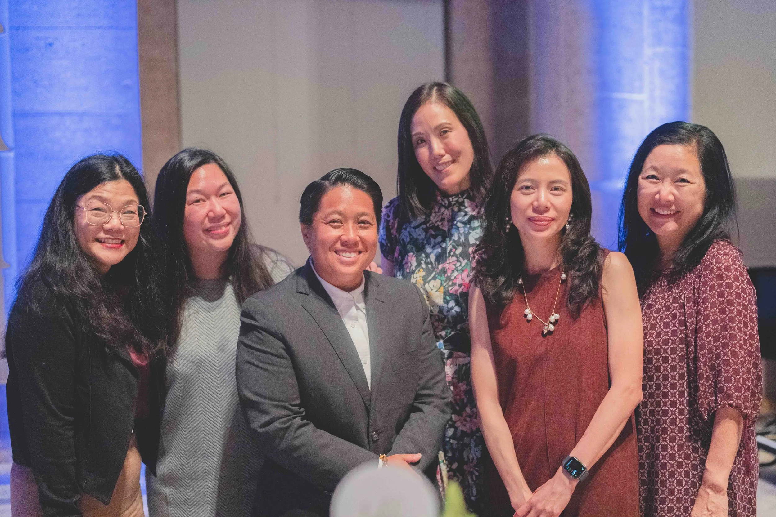 Group of six diverse professional women and one man, smiling and posing together indoors, dressed in business casual and formal attire.