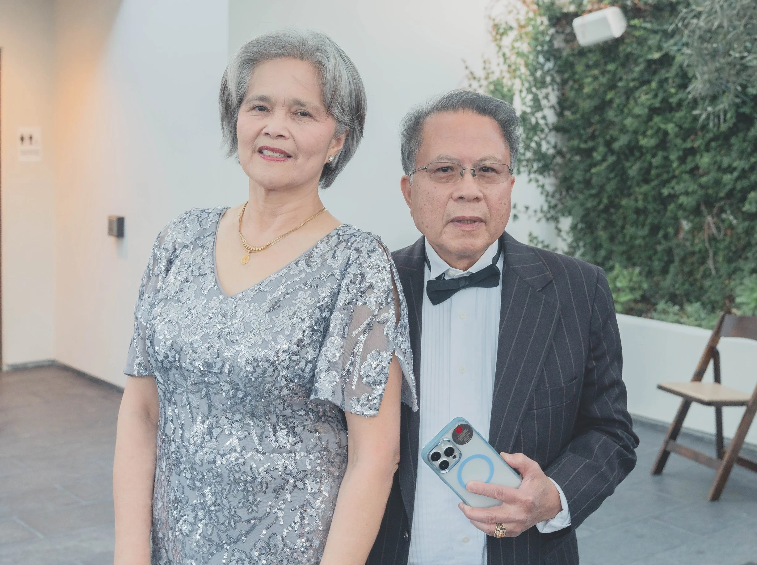 An elegantly dressed couple, standing together, with the man holding a smartphone. The woman is wearing a silver lace dress, and the man is in a suit and bow tie.