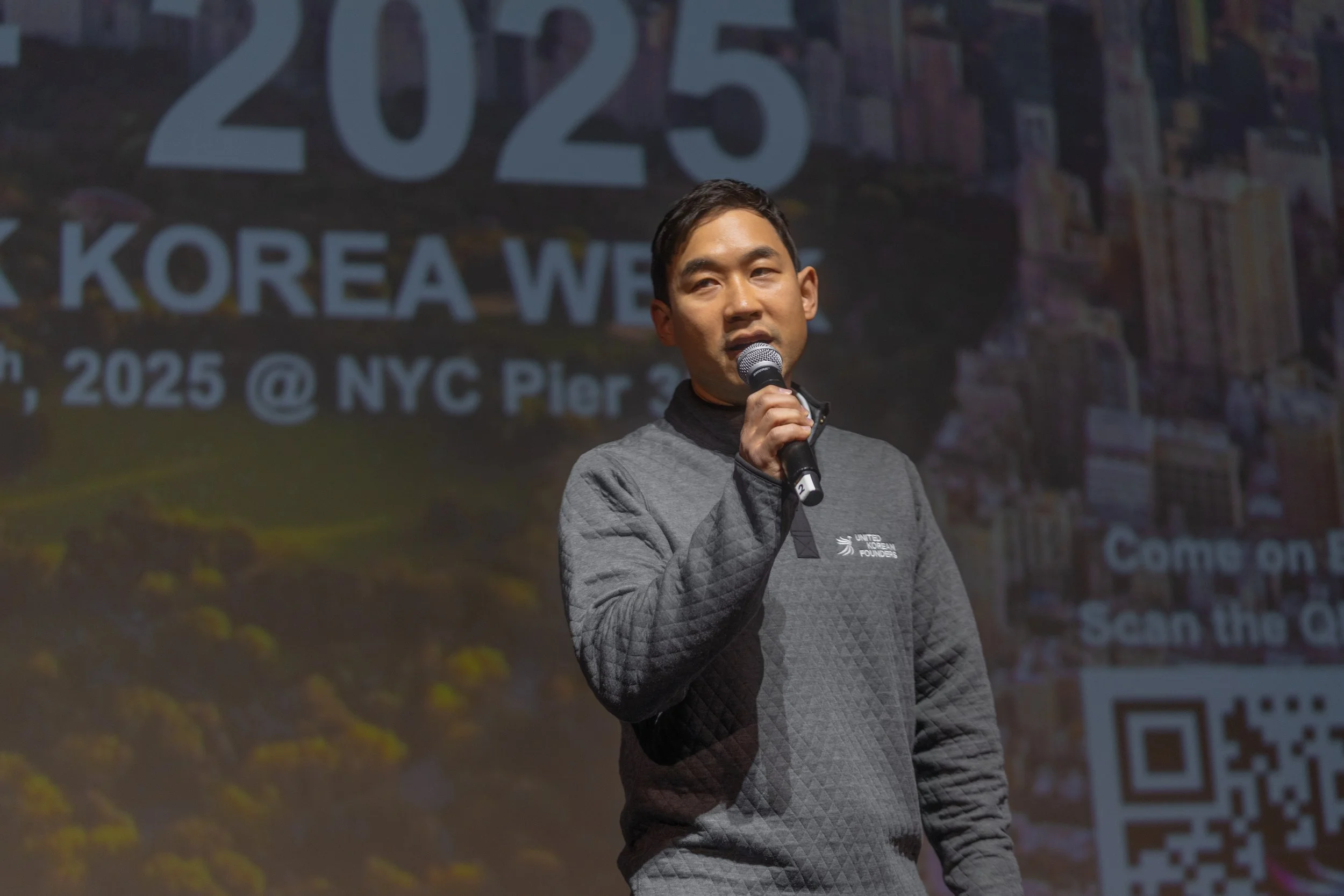Person speaking on stage with microphone, wearing a quilted sweater, against a background announcing "Korea Week 2025" event in NYC.