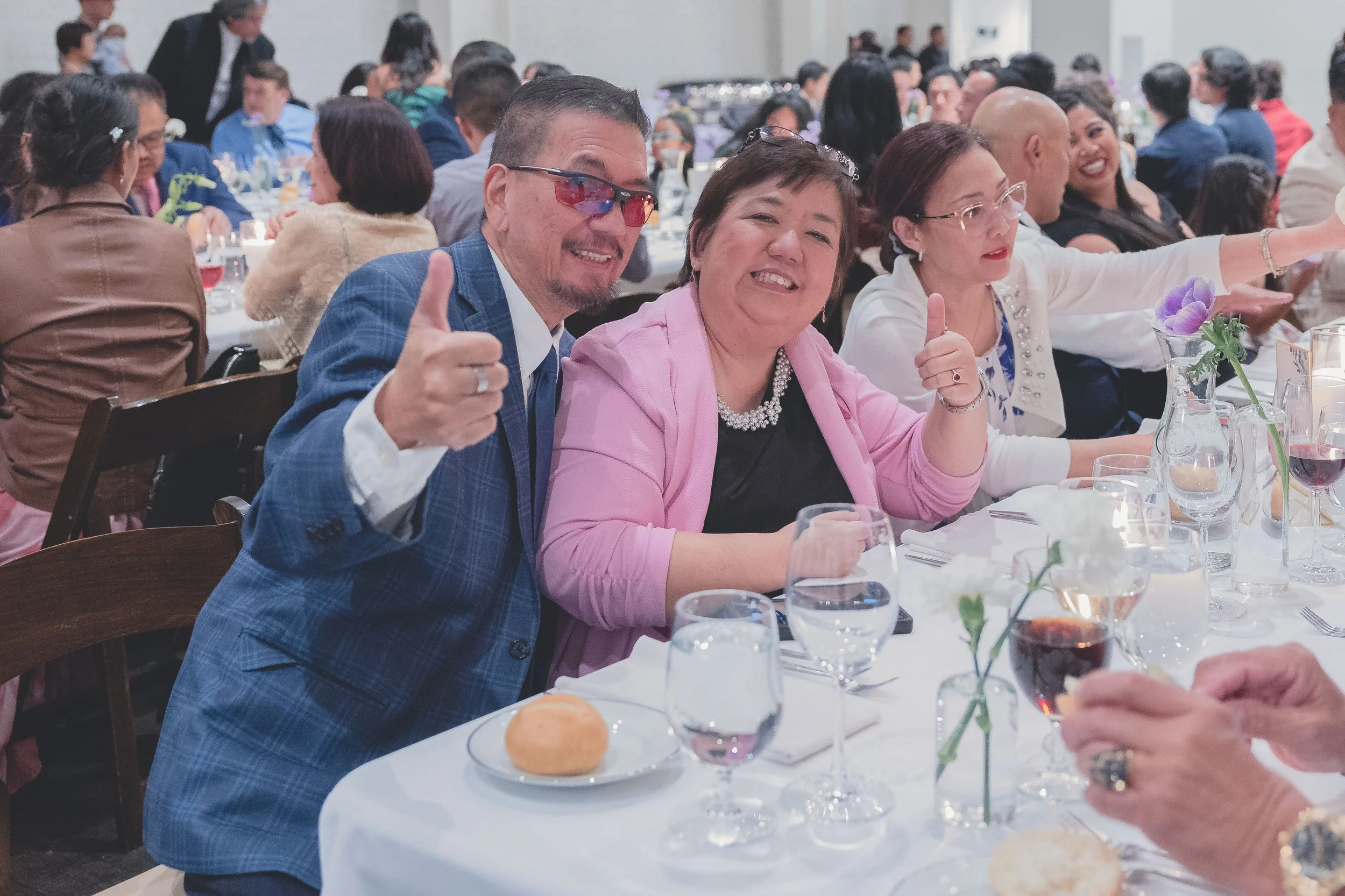 Guests at a formal dinner event sitting at a table, smiling and gesturing with thumbs up, with glasses and a bread roll on the table.