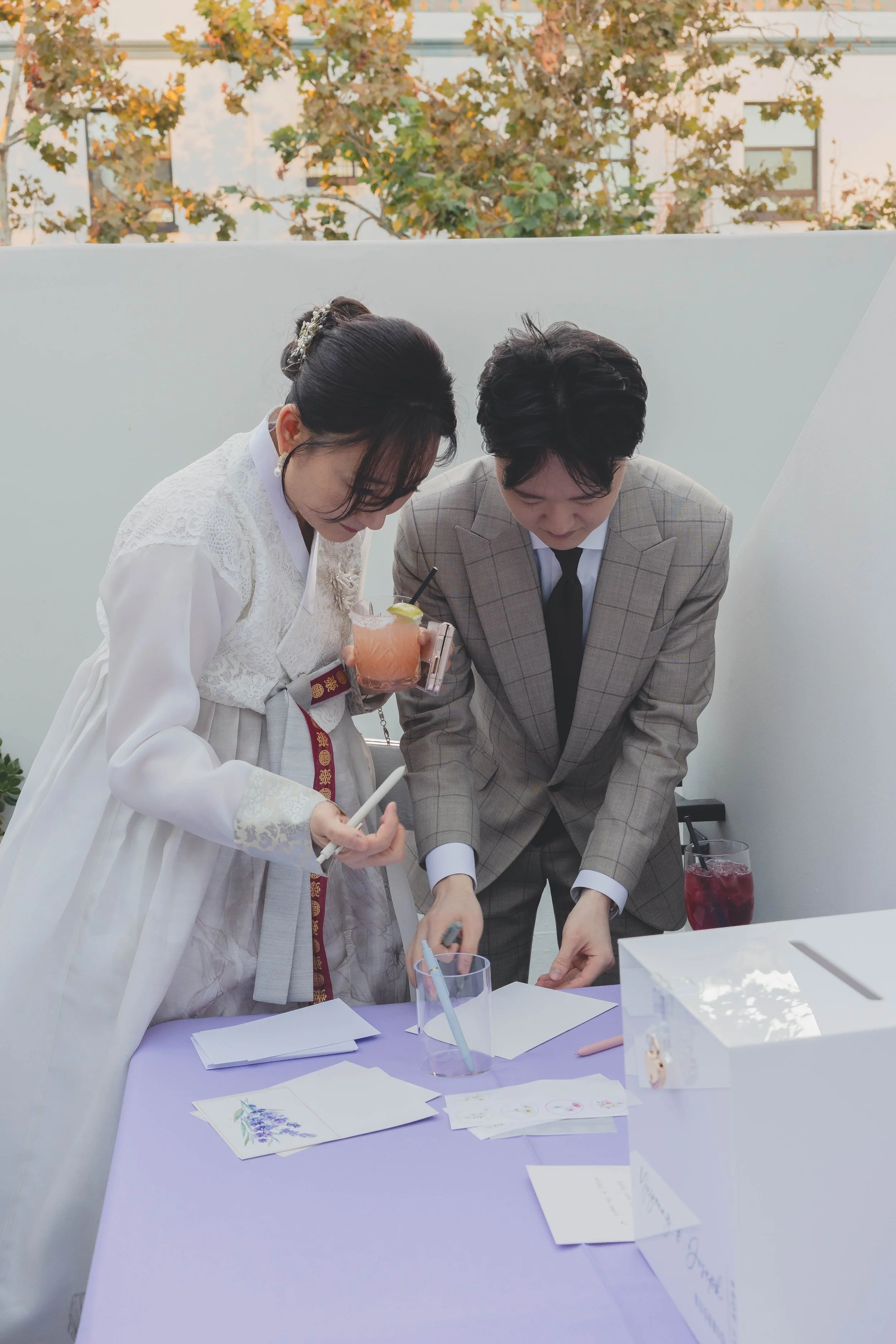 A woman in traditional Korean hanbok and a man in a gray suit fill out papers at a table with a ballot box. They are outdoors, with trees in the background.