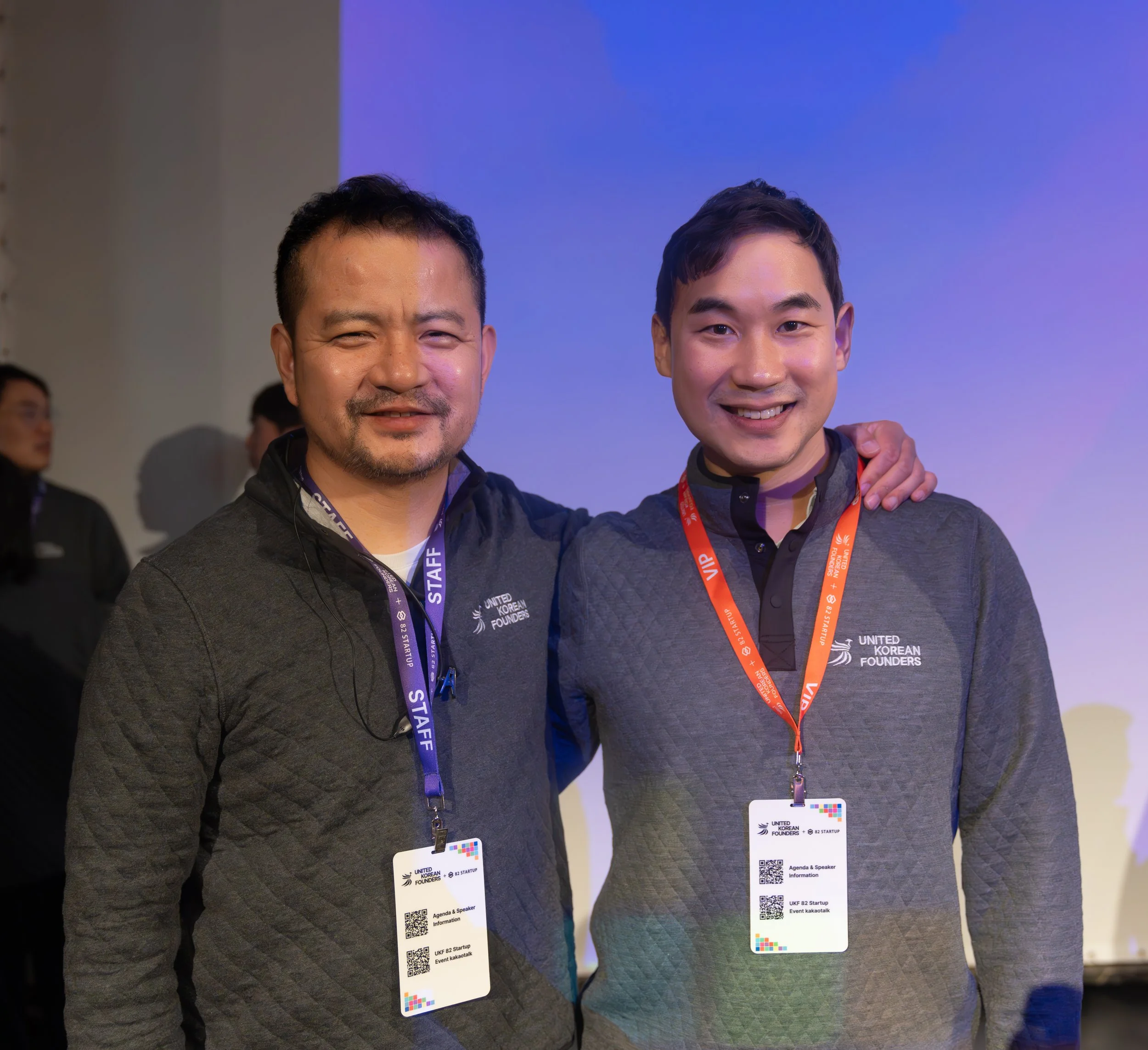 Two men smiling with event badges and United Korean Founders shirts.