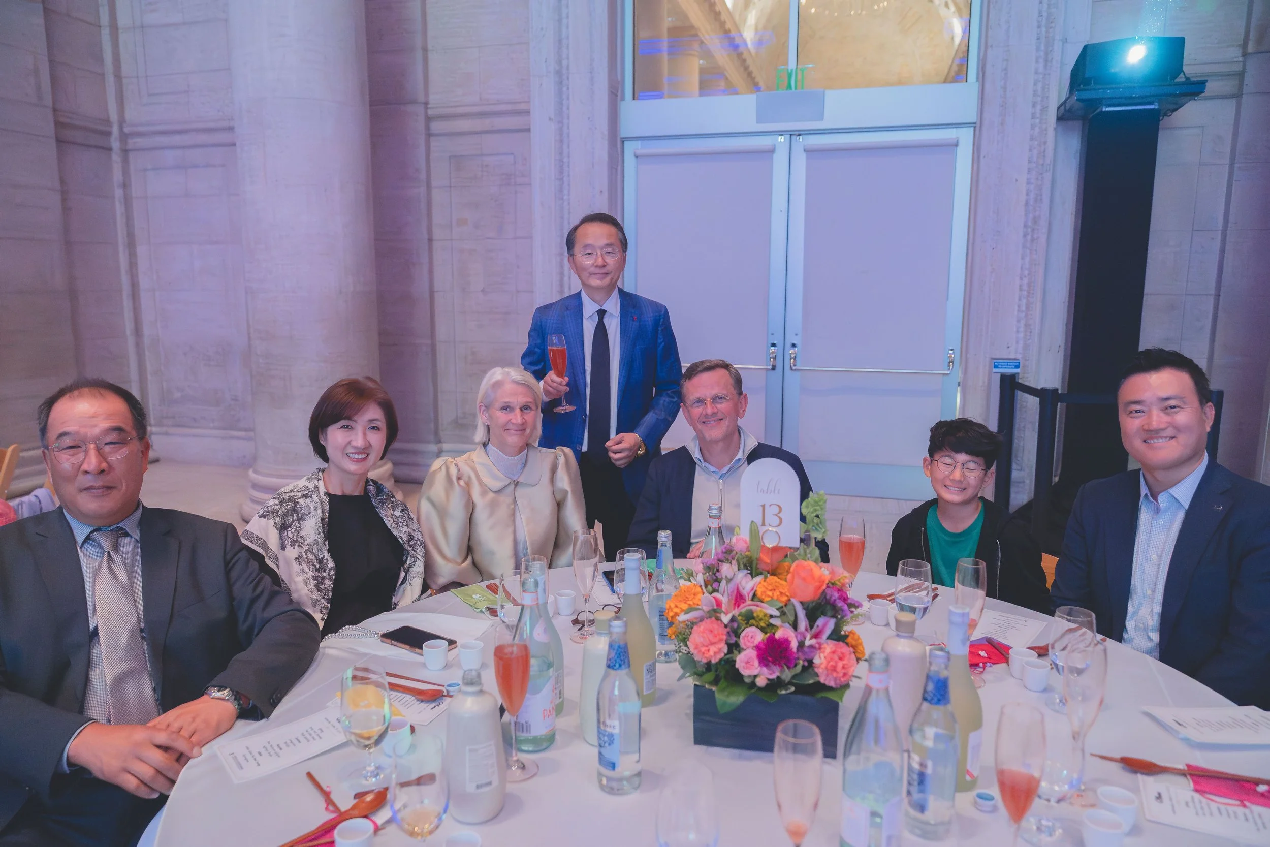 A group of seven people, six sitting and one standing, at a formal dinner event. The table has floral centerpiece, drinks, and menus. The setting appears to be an elegant venue with stone walls and high ceilings.