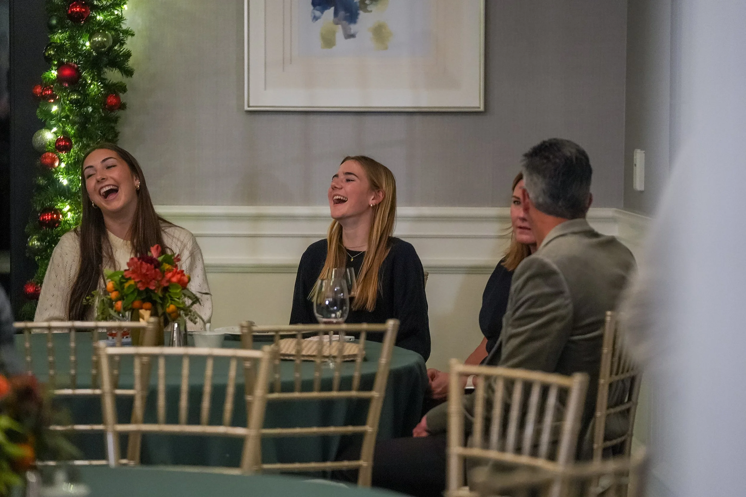 Group of people sitting at a table with holiday decorations, laughing and talking, in a warmly lit room.