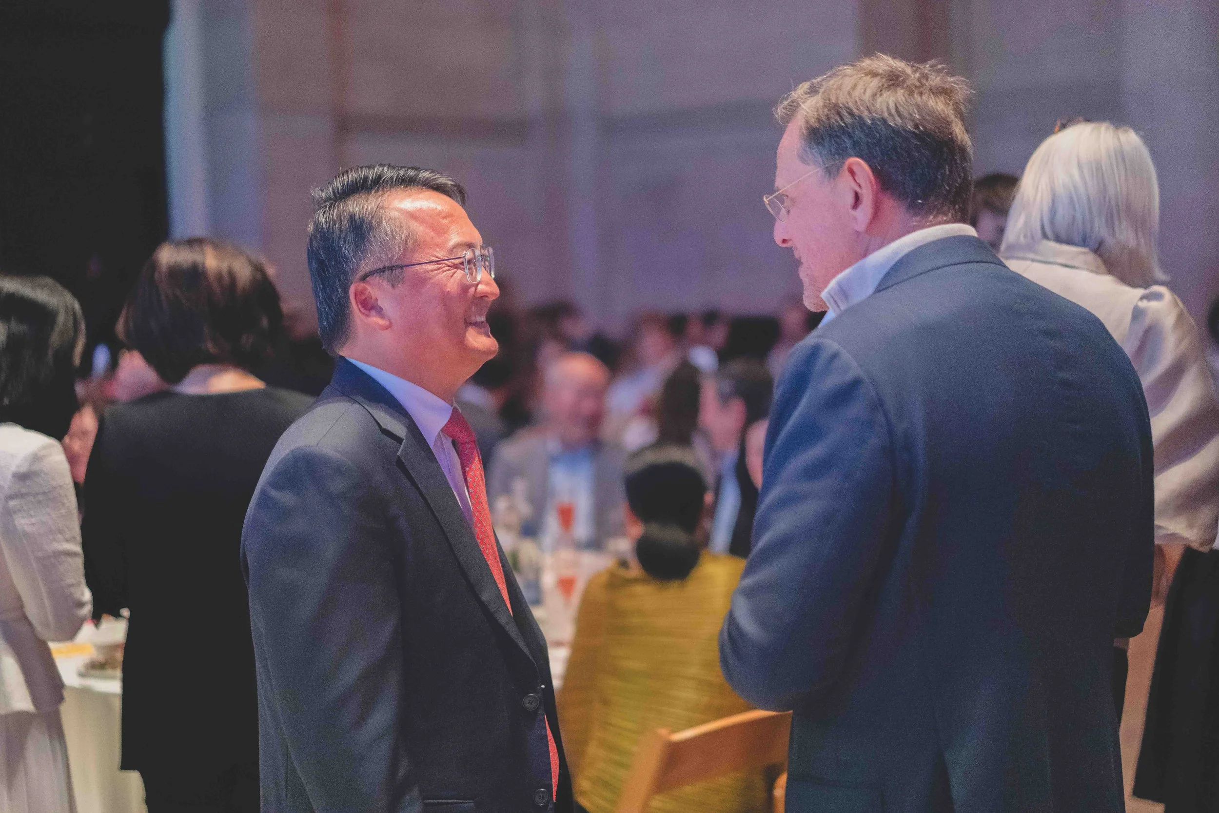 Two men in suits are smiling and talking at a social event in a large banquet room with many people in the background.