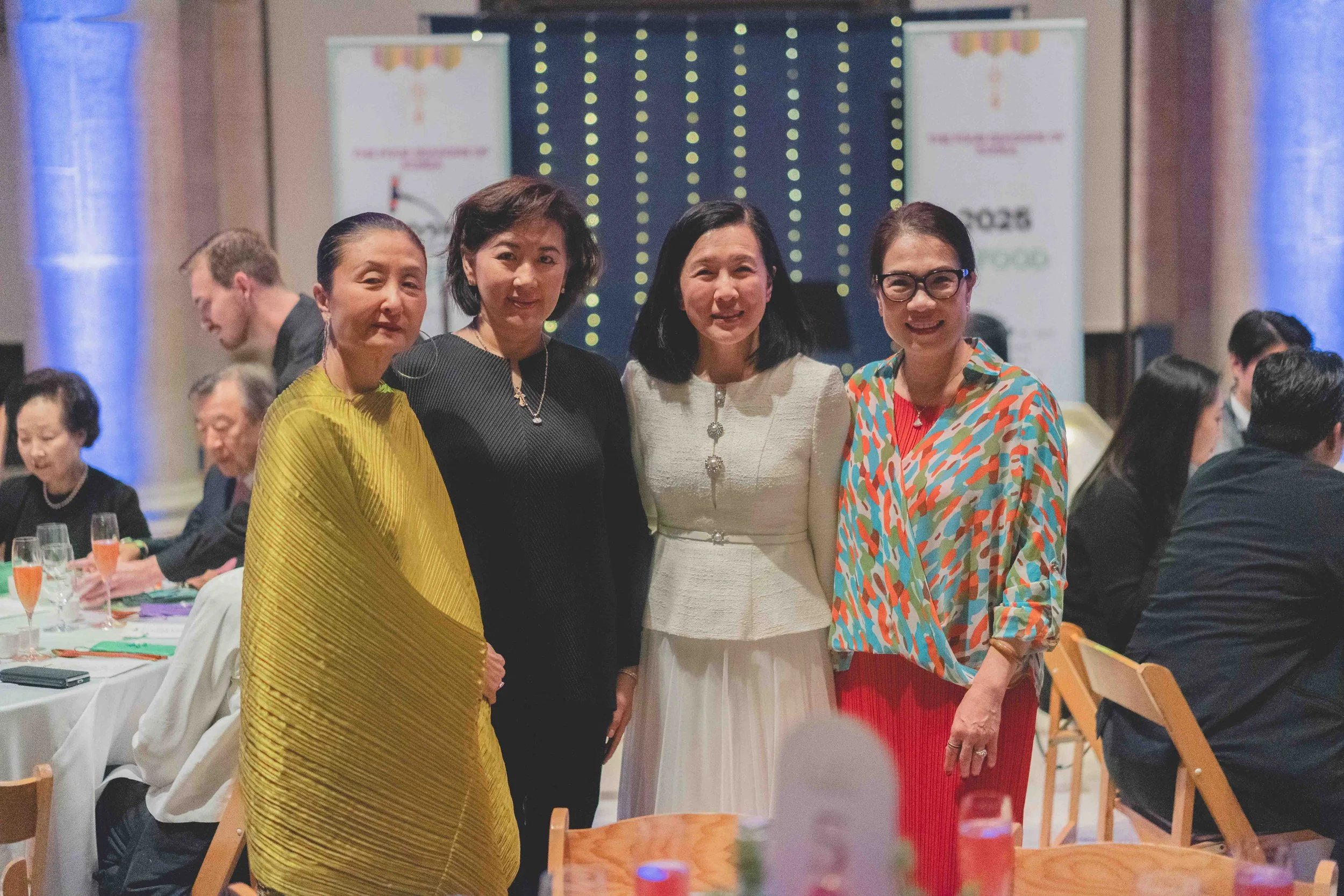 Four women standing together at a formal event, smiling for the camera, with tables and other attendees in the background.
