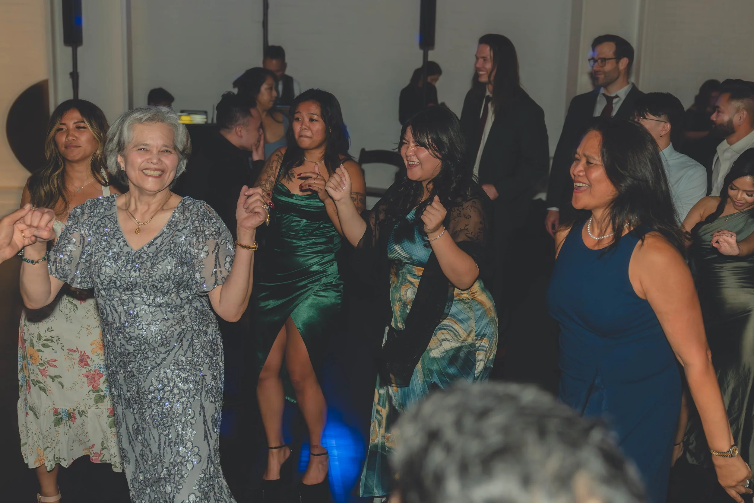 A group of people dancing and smiling at a formal event. They are dressed in elegant attire, with women in dresses and a man in a suit. The atmosphere appears lively and festive.