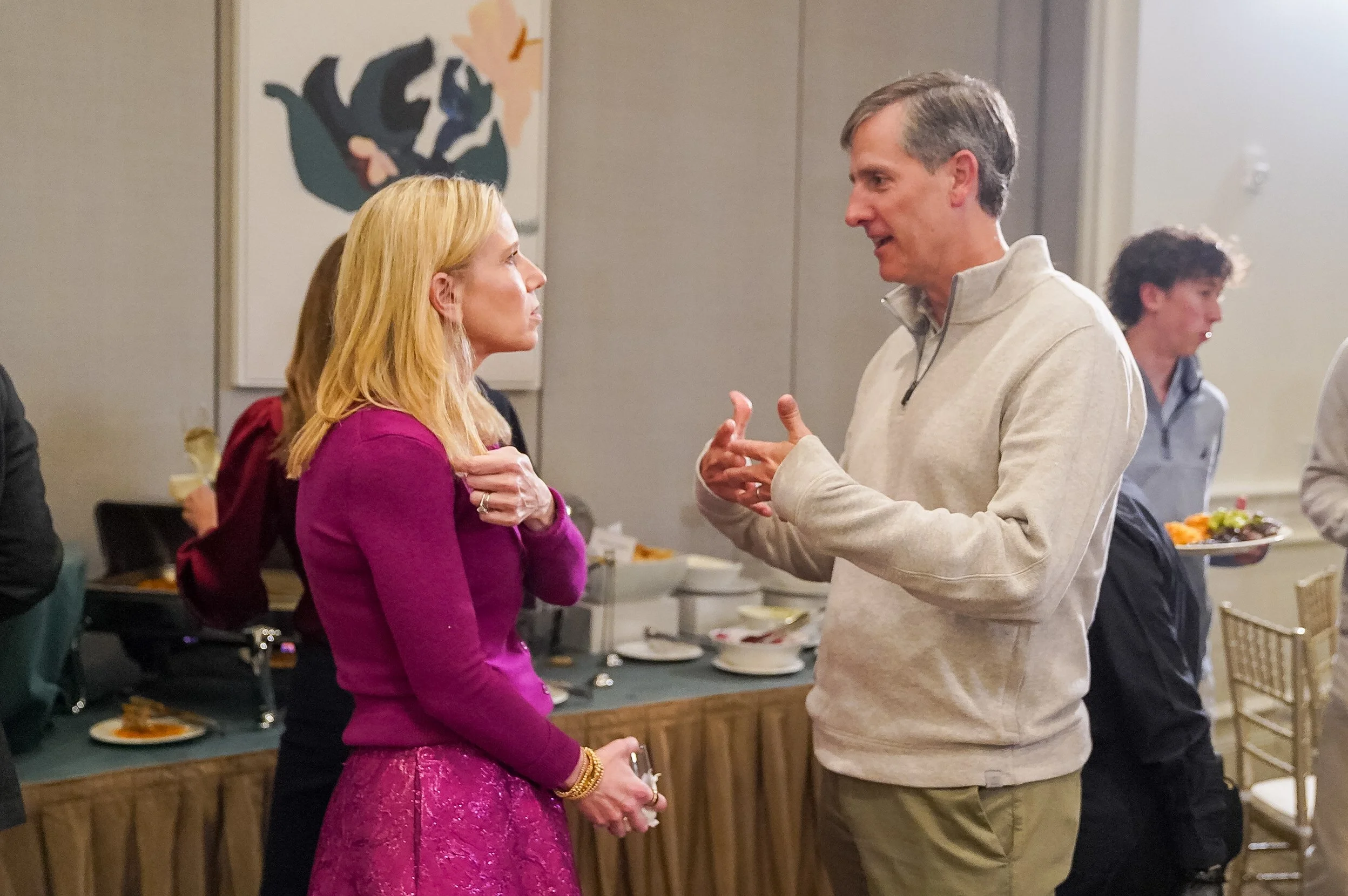 Two people having a conversation at a social gathering with food and drinks, while a man in the background carries a plate of food.