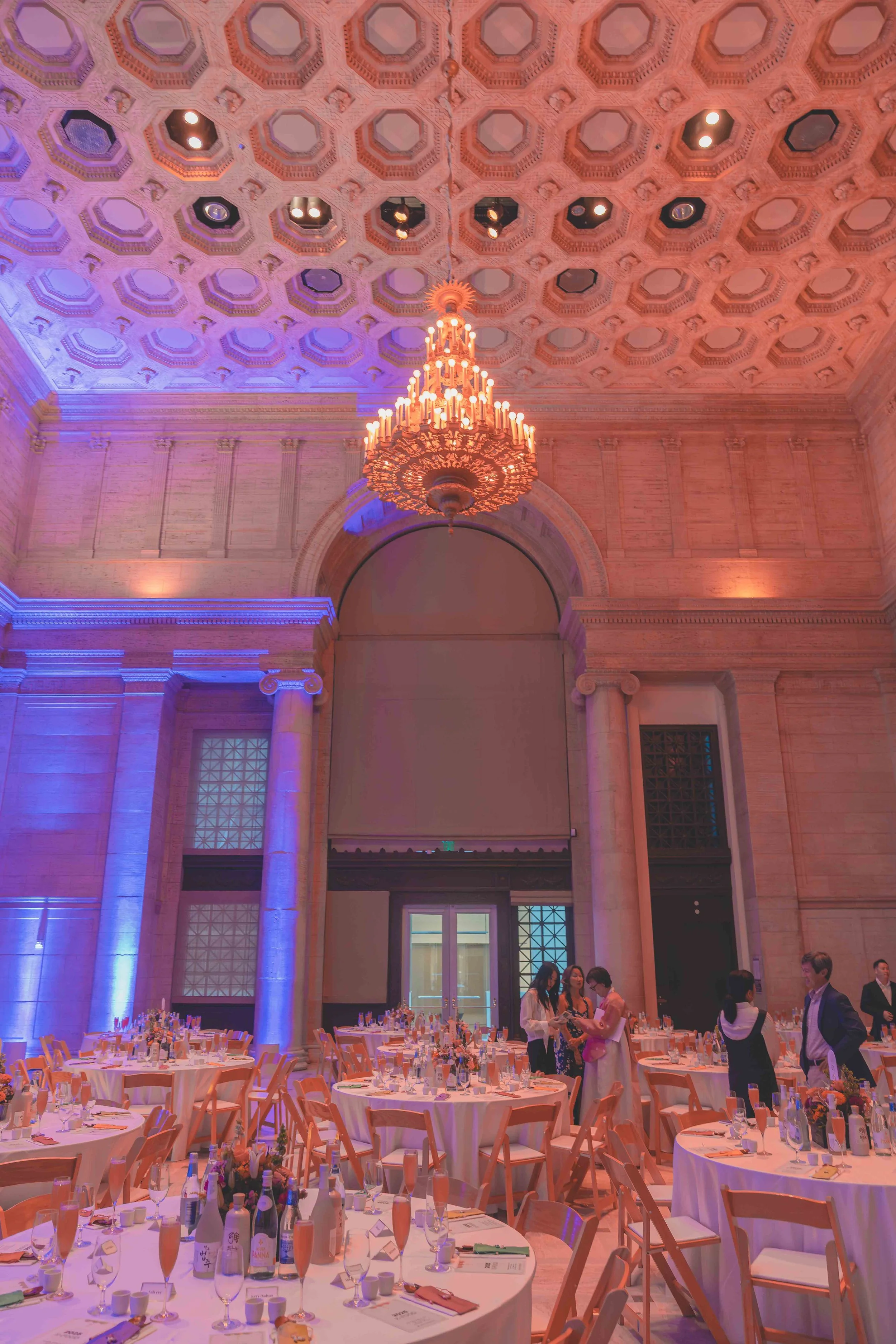 Elegant banquet hall decorated for an event with round tables set with glassware, napkins, and floral centerpieces, and people standing and conversing near the tables. The hall has a high ornate ceiling with a large chandelier and colorful lighting.