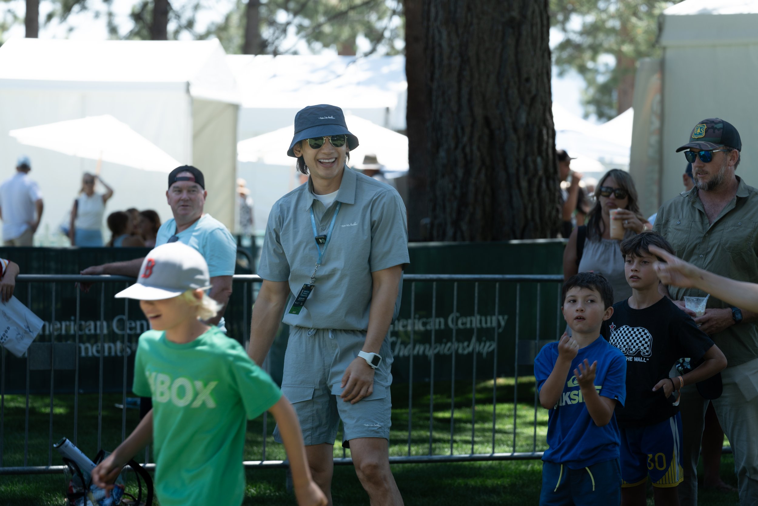 Group of people outdoors at a public event, including a man in a bucket hat and sunglasses, with several tents and trees in the background.