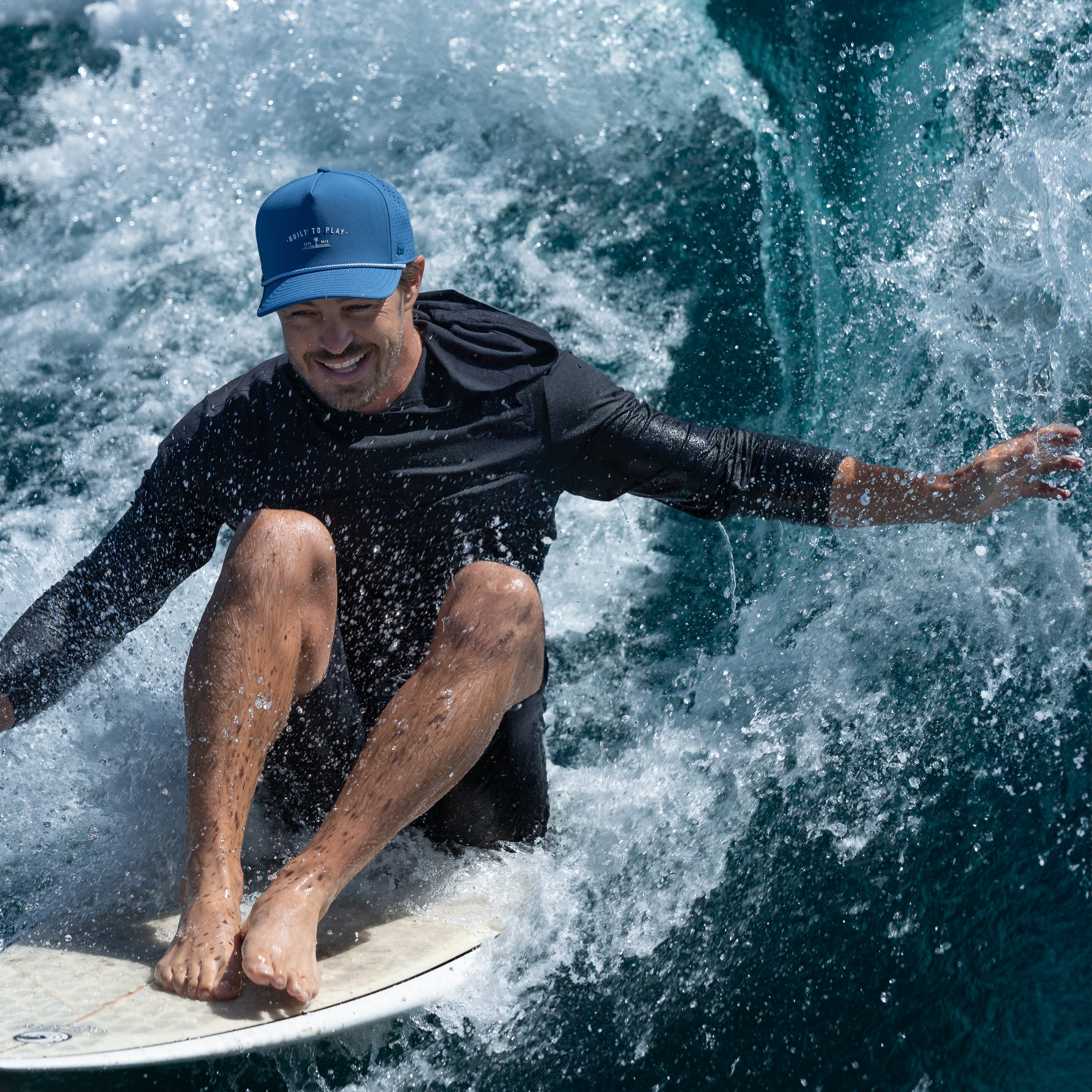 Surfer riding a wave on a surfboard, wearing a blue cap and dark long-sleeve shirt, smiling amidst splashing water.