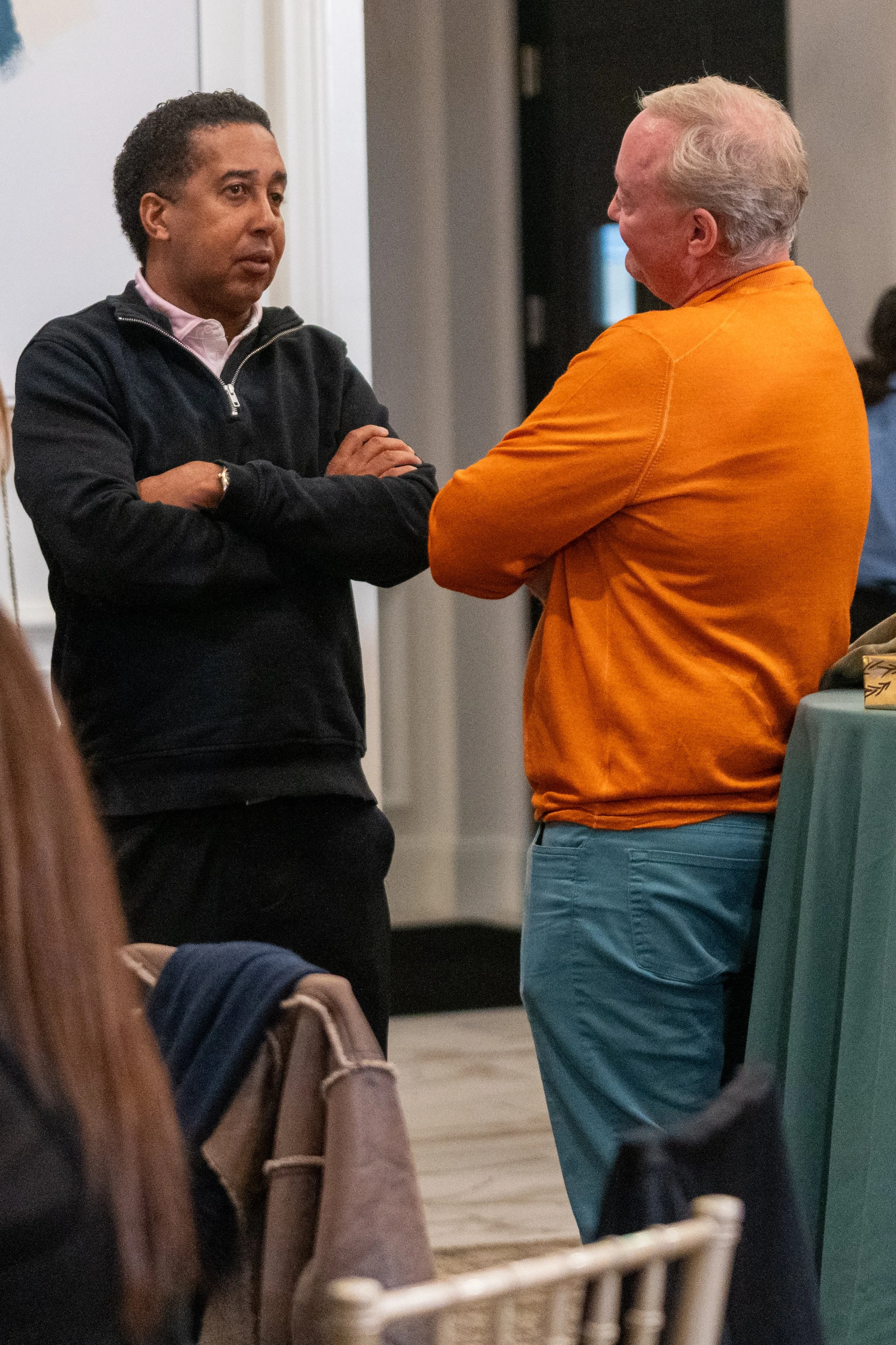 Two men having a conversation in a room with one wearing a dark sweater and the other in an orange sweater.
