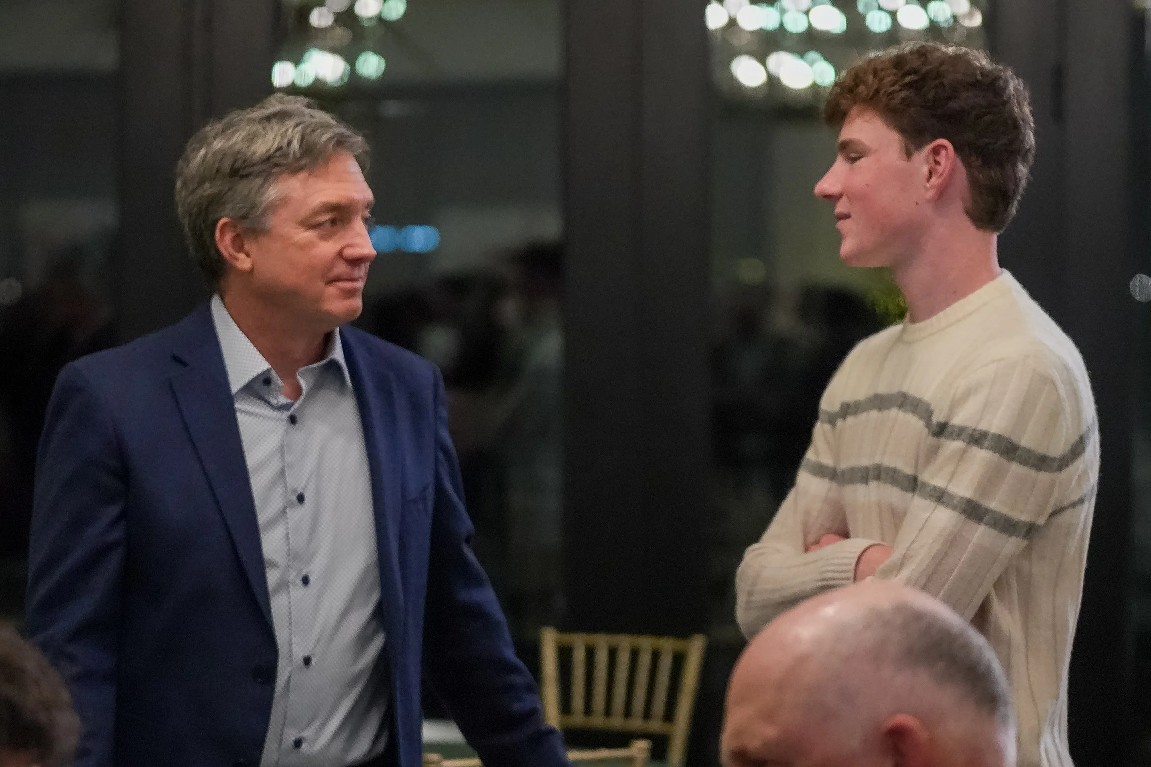 Two men having a conversation indoors, one in a blue suit and the other in a striped sweater.