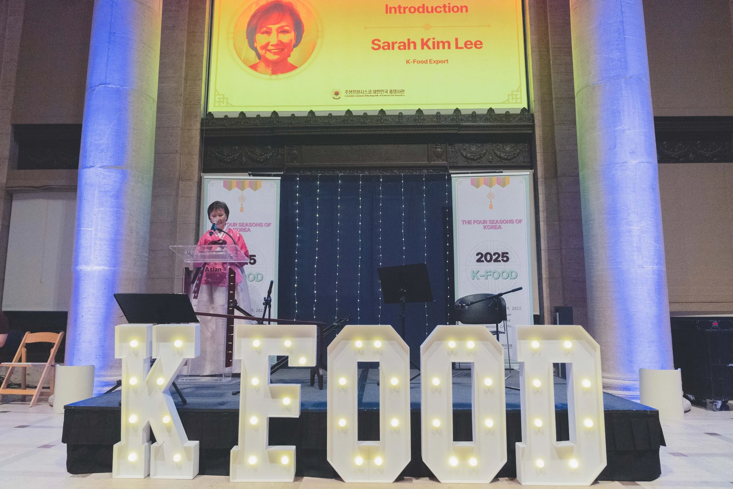 A woman standing at a lectern on a stage decorated with large illuminated letters spelling 'KE100', with a large projection screen behind her displaying her name, Sarah Kim Lee, and a portrait of her. The stage also has two banners with Korean text a