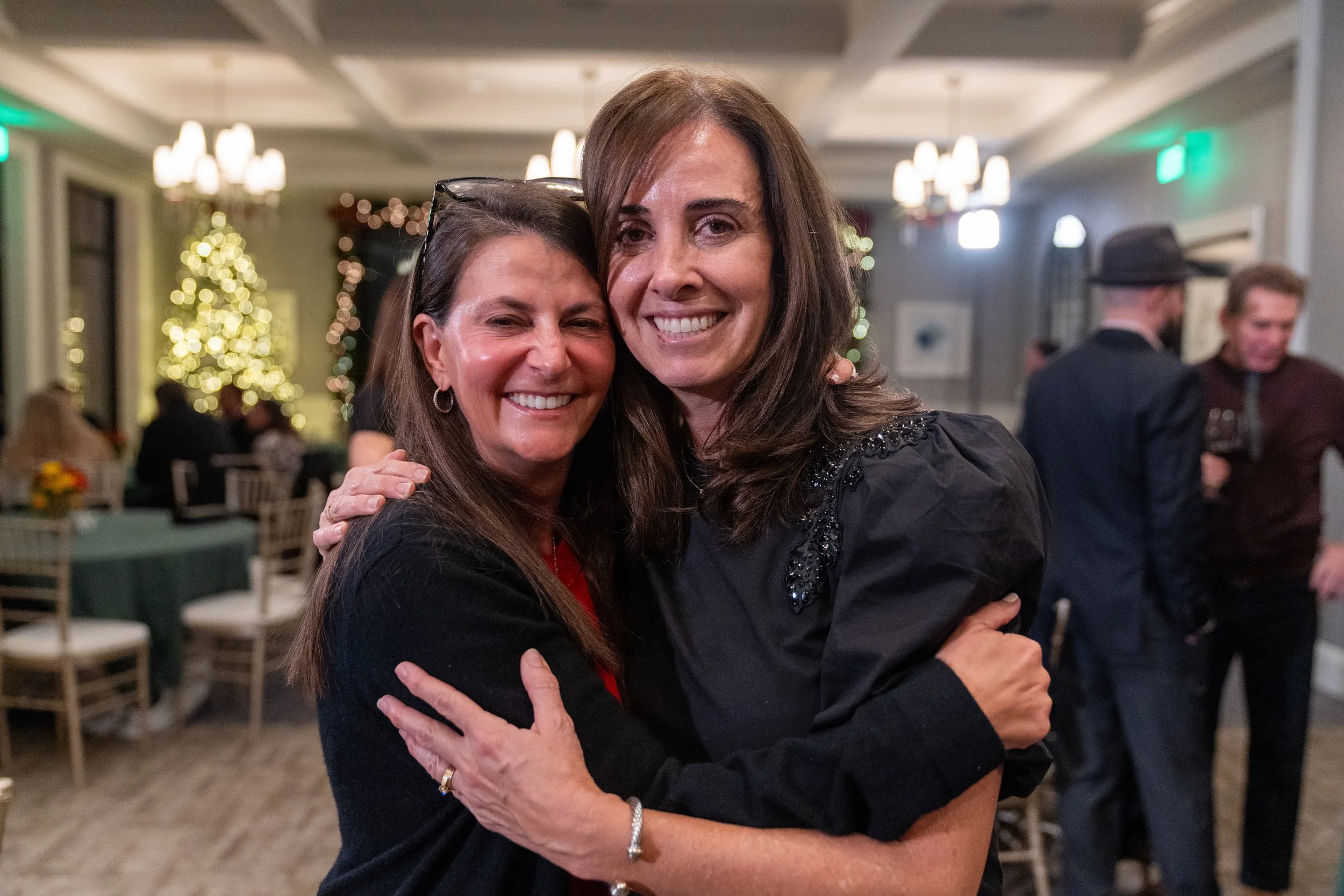 Two women hugging and smiling at a social gathering with holiday decorations in the background, including a lit Christmas tree.
