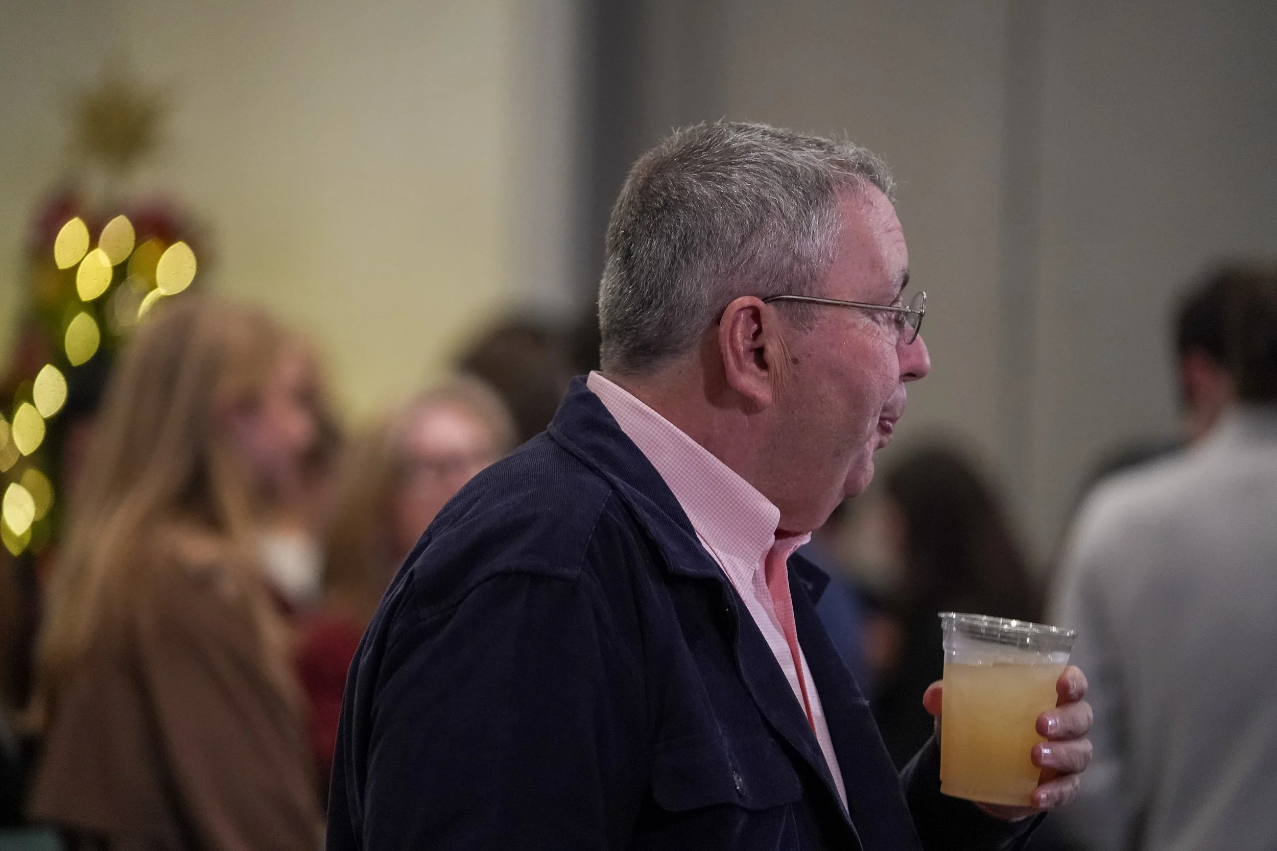 Man holding a drink in a social gathering with blurred people in the background.