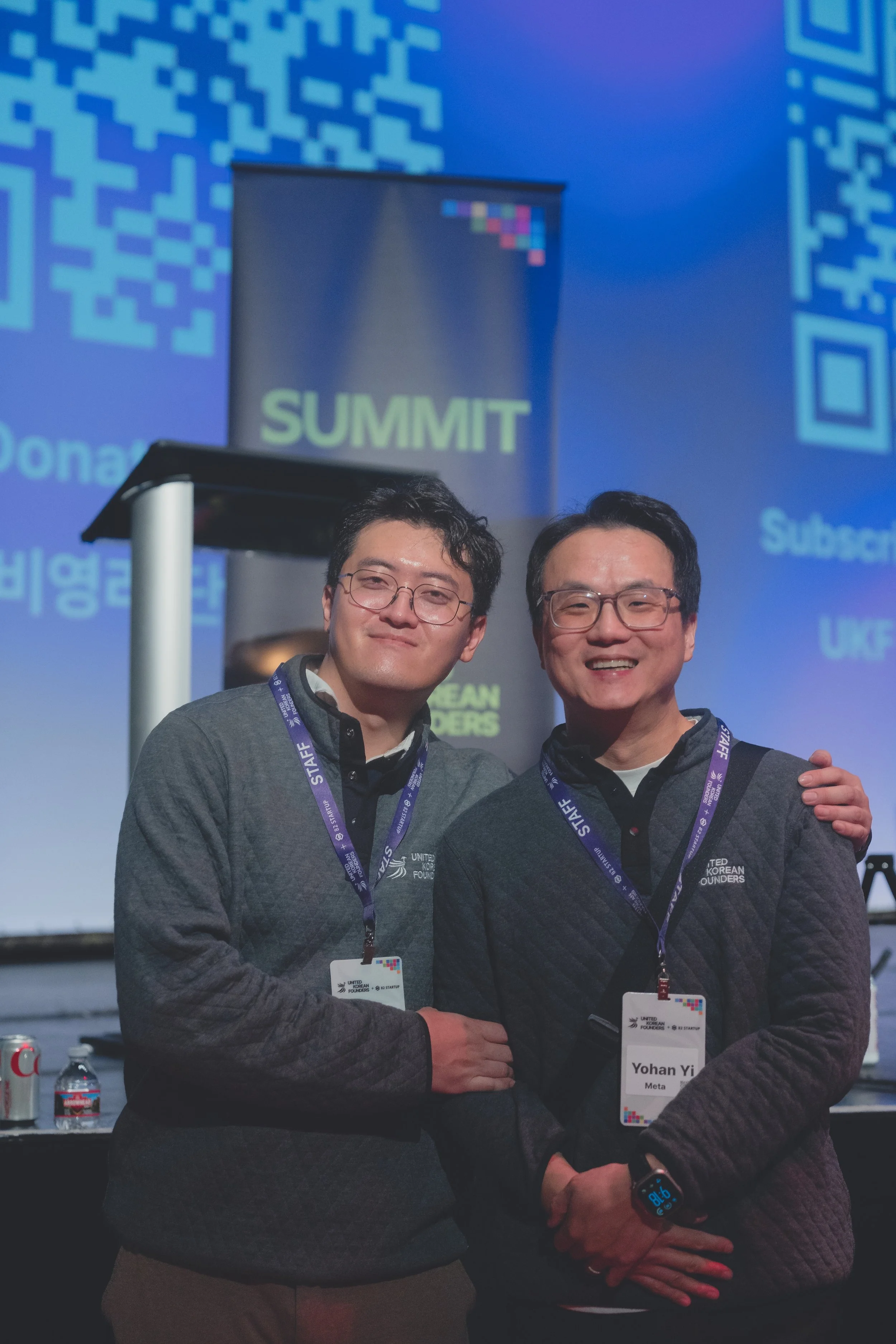 Two people wearing conference badges and staff lanyards at a summit. They are smiling and standing in front of a blue screen with QR codes. A banner with ‘SUMMIT’ is visible.