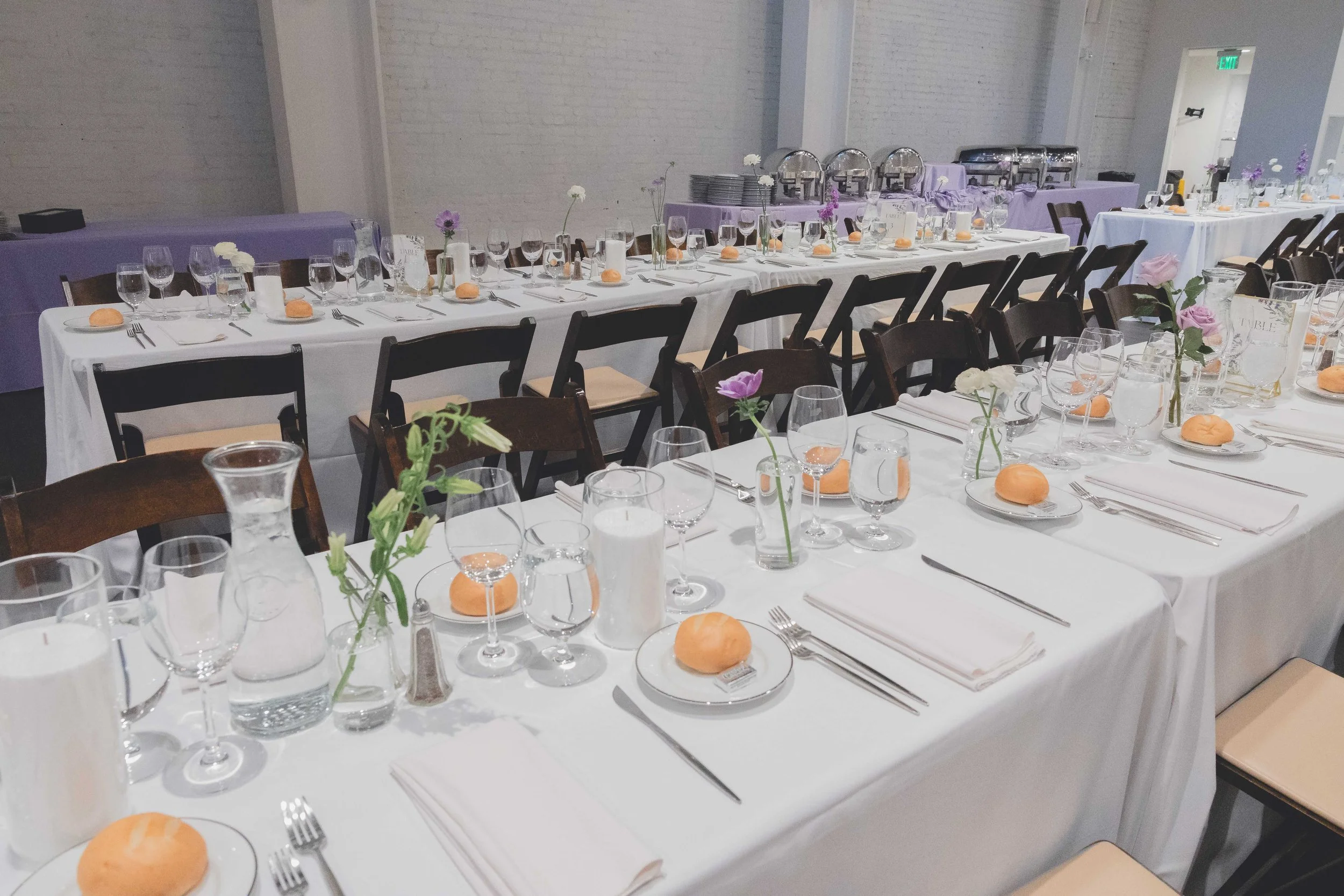 Elegant banquet hall setup with long tables dressed in white tablecloths, neatly arranged tableware, bread rolls on plates, glassware, and minimal floral centerpieces; wooden chairs are placed around the tables.