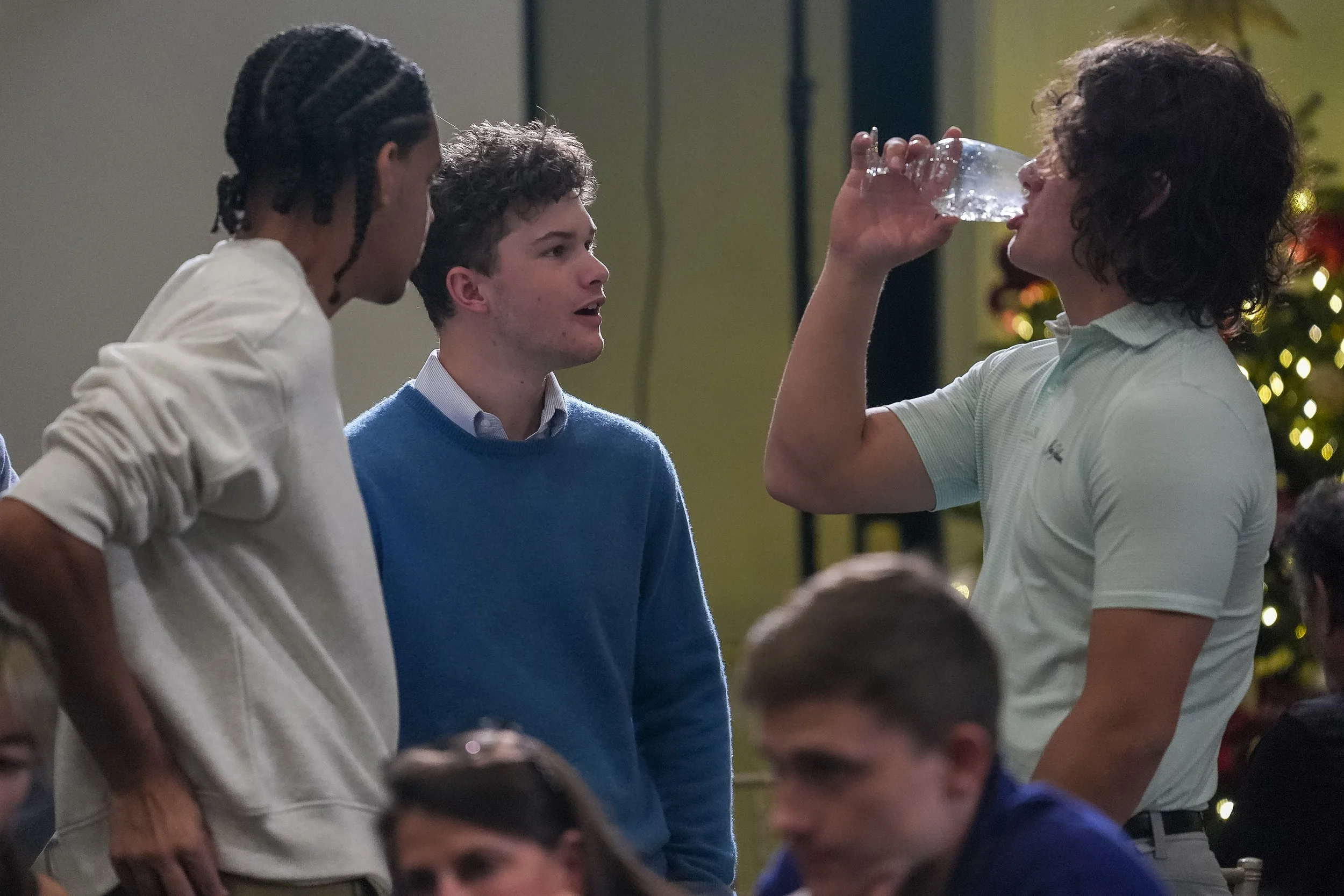 Three young men in casual attire conversing, one drinking from a plastic cup, in a social setting.