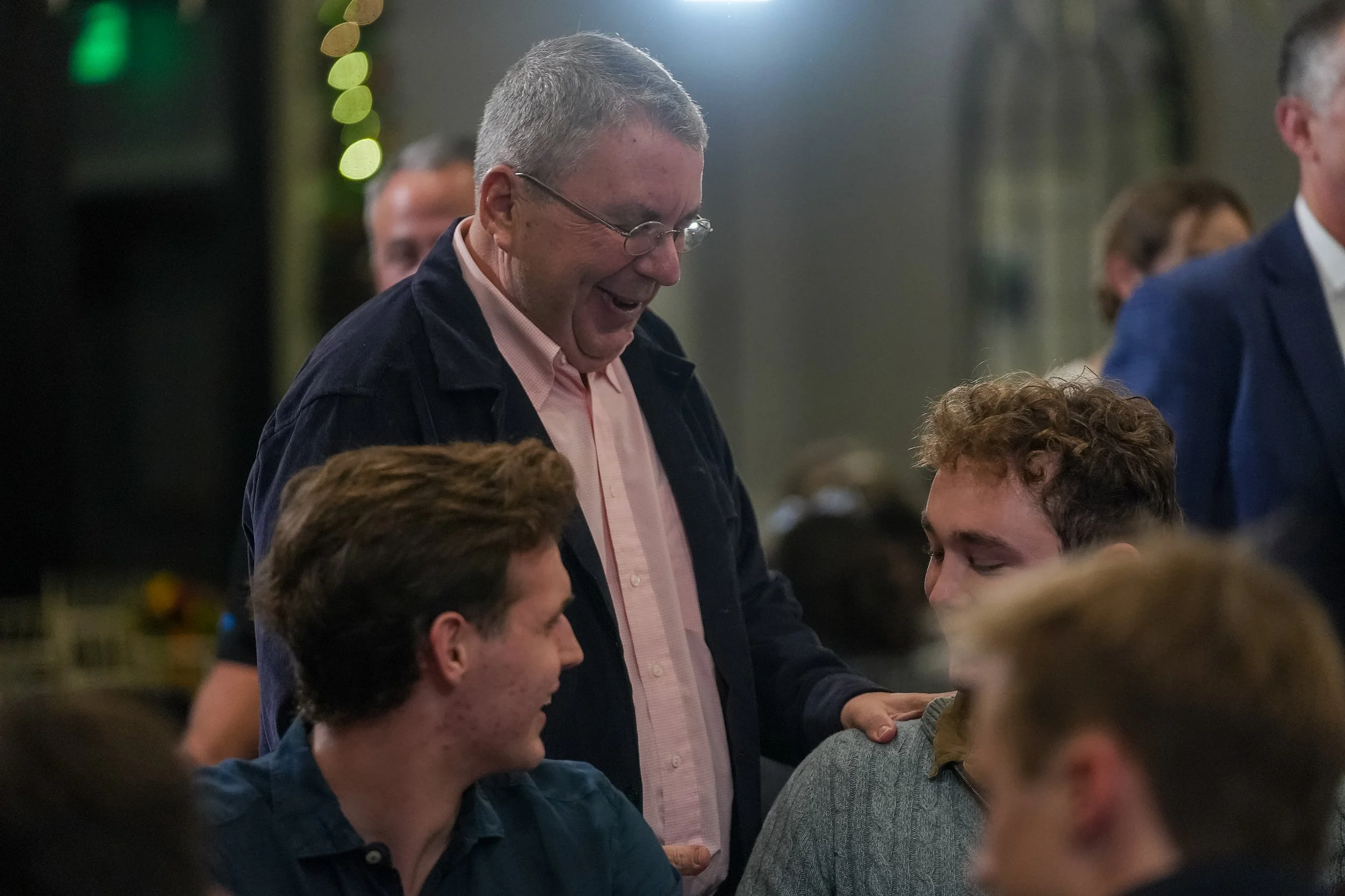 A man in glasses and a dark jacket smiles while interacting with seated young men at a gathering. The scene is indoors with soft lighting.