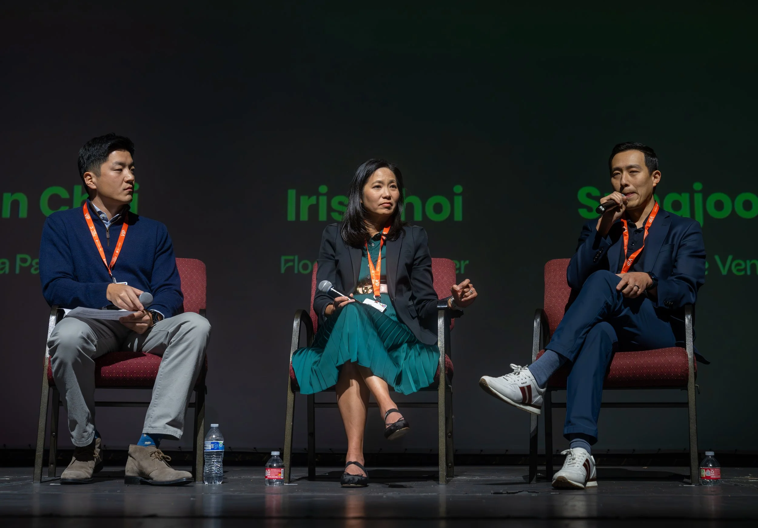 Panel discussion with three people seated, each holding microphones, wearing professional attire, in front of a dark background with green text.