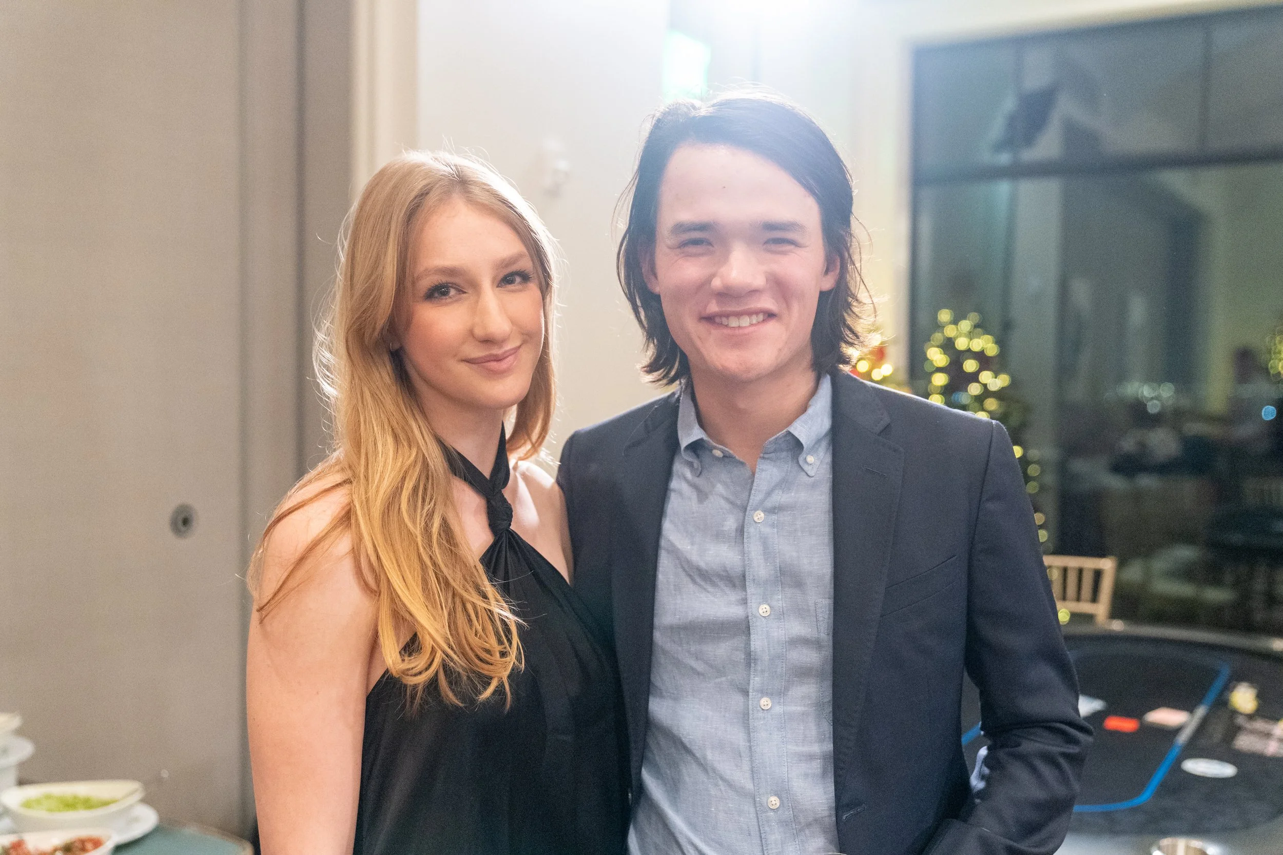 A young woman with long blonde hair wearing a black dress and a young man with long dark hair wearing a light blue shirt and dark blazer stand smiling together in a room decorated with lights.