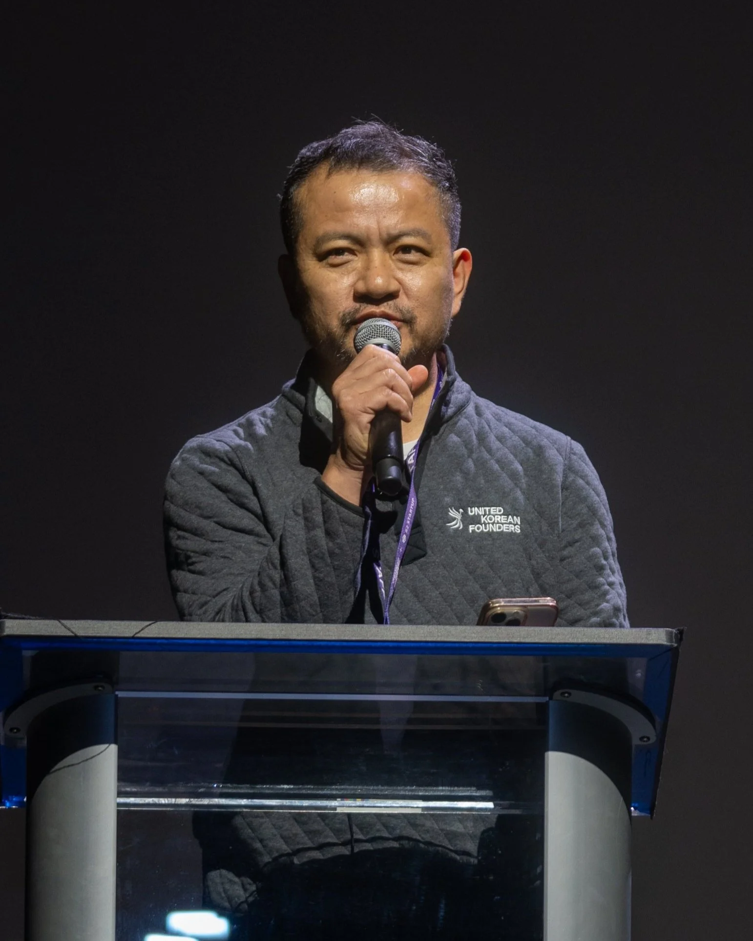 A man speaking into a microphone at a podium, wearing a jacket with "United Korean Founders" embroidered on it.